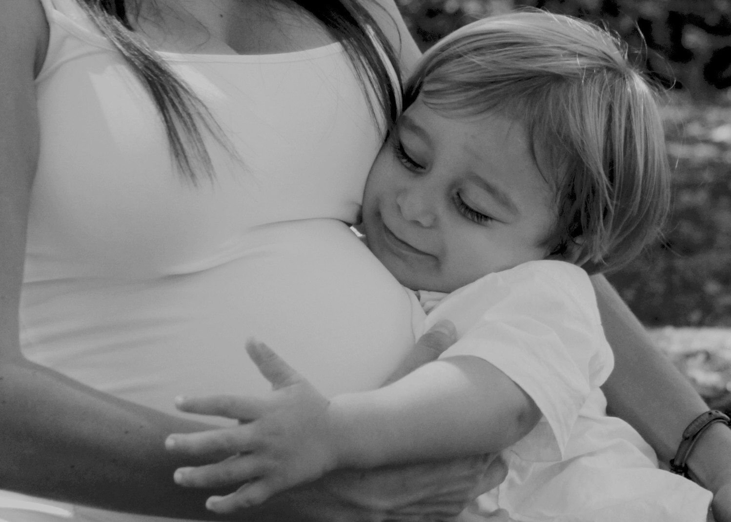 Black and white close-up of a young child hugging and kissing a pregnant mothers belly outdoors.