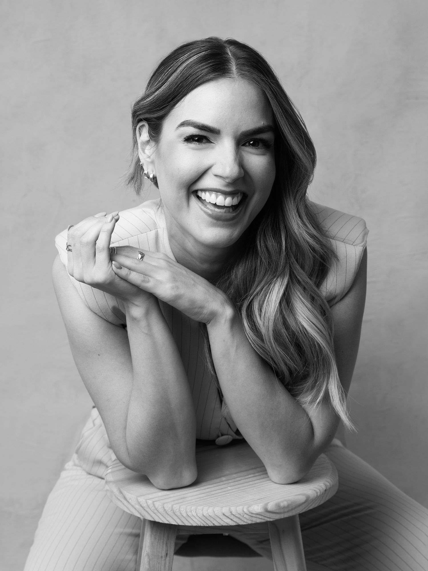Black and white studio portrait of laughing woman with wavy hair leaning on stool.