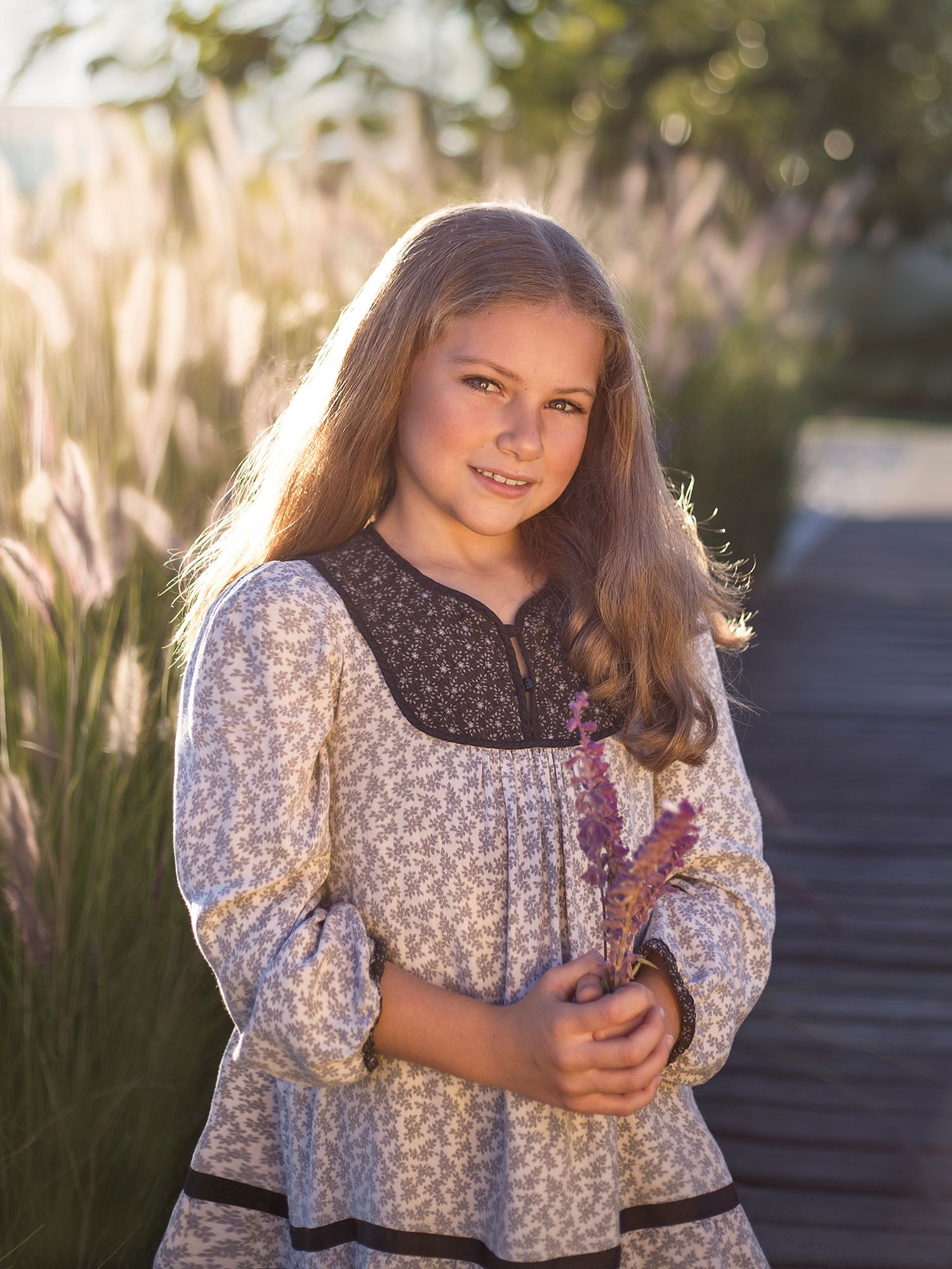 Young girl in floral dress holding lavender flowers in warm golden sunset outdoor portrait.