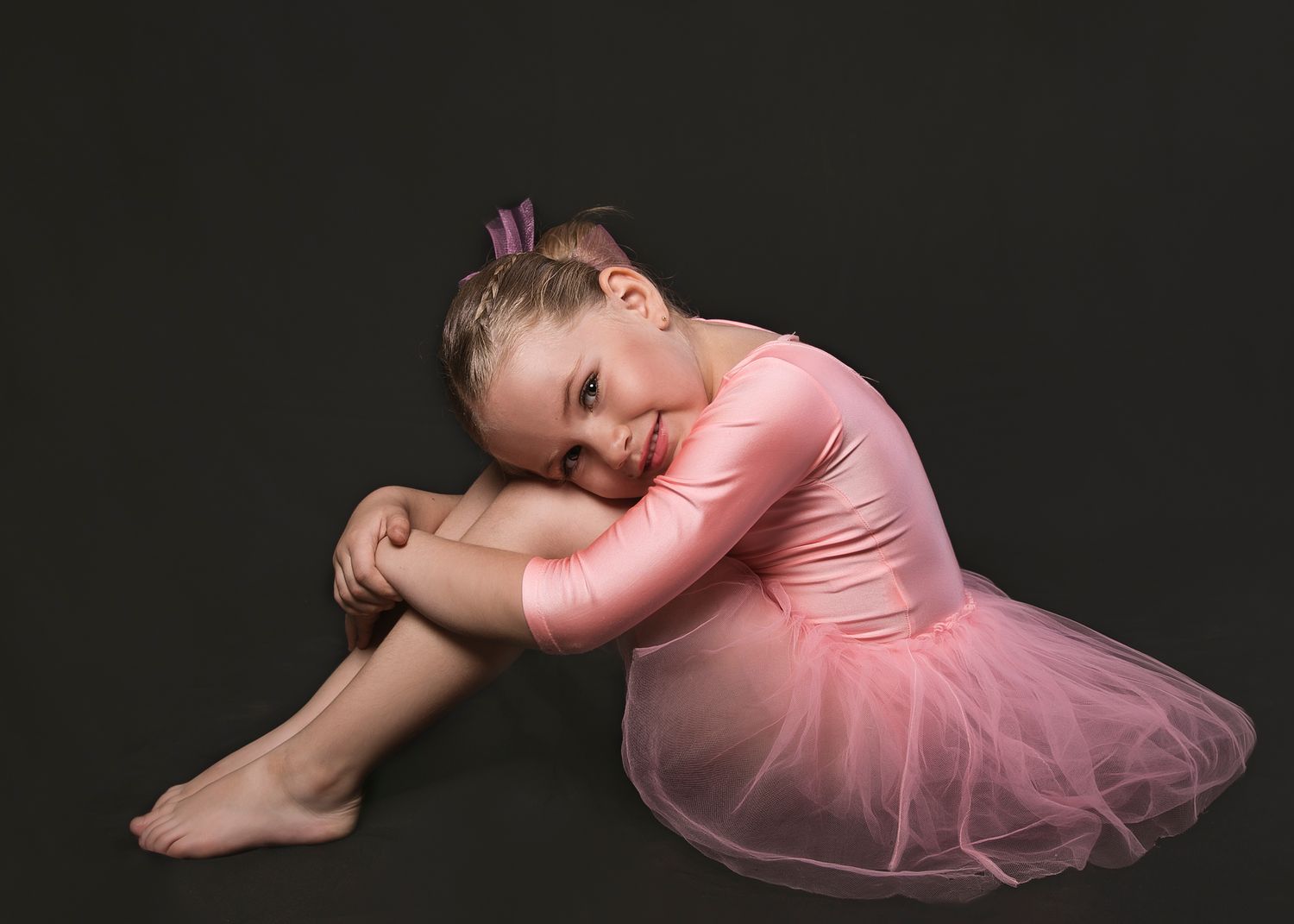 Young girl in a pink ballet tutu and leotard sitting gracefully against a dark background.