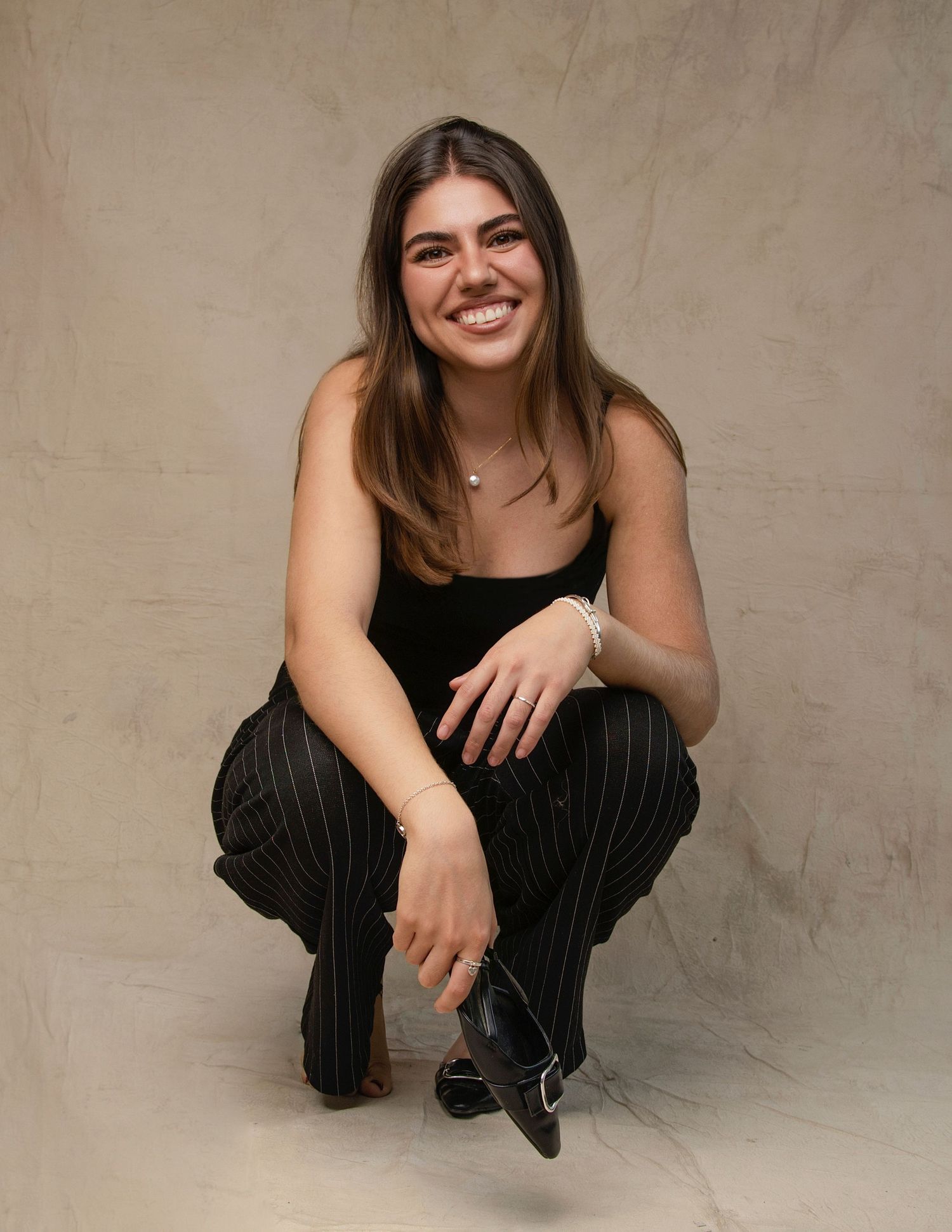 Young woman in black top and pinstripe pants smiling while crouching in studio portrait session.