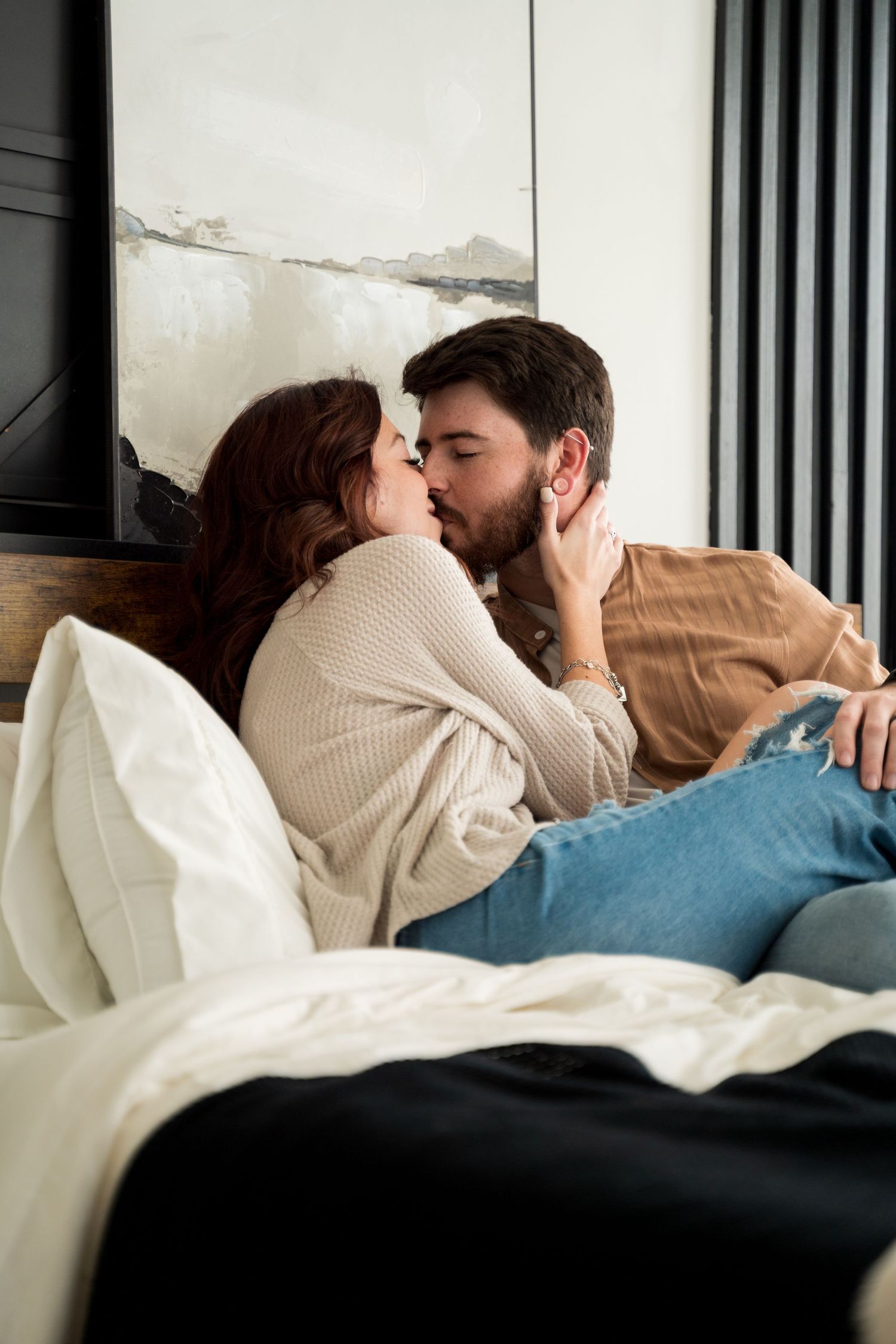 A couple sharing a romantic kiss while sitting together on a bed, dressed in casual clothing.