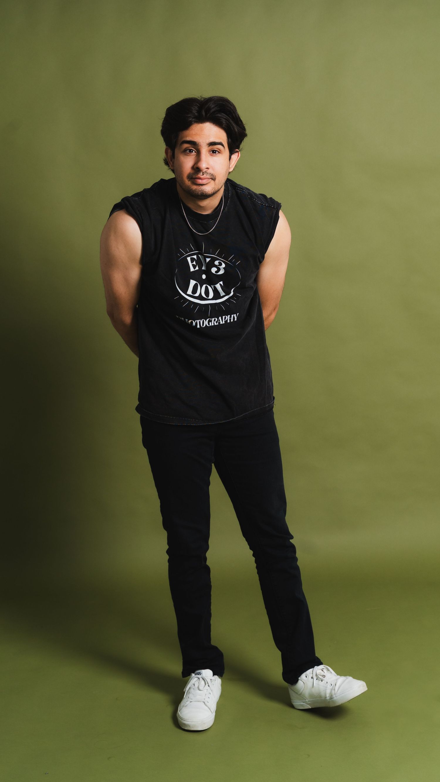 Young man in black sleeveless shirt and jeans posing confidently against an olive green studio background.