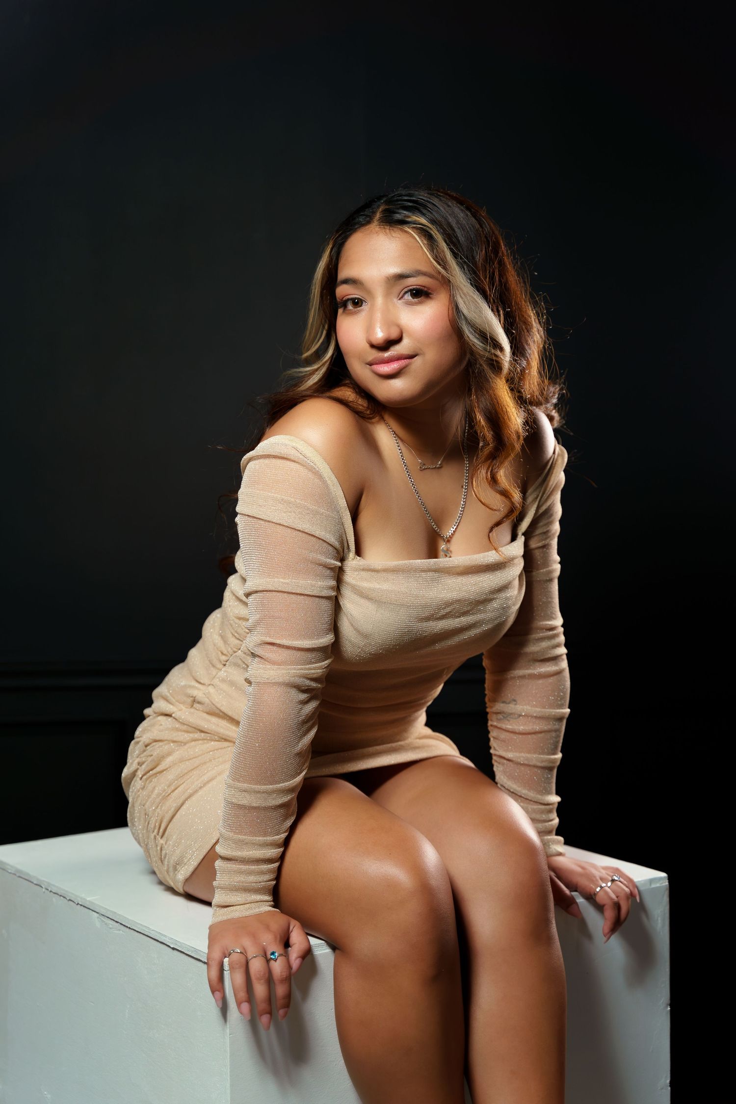 Young woman in beige off-shoulder mini dress seated on white cube against dark background.