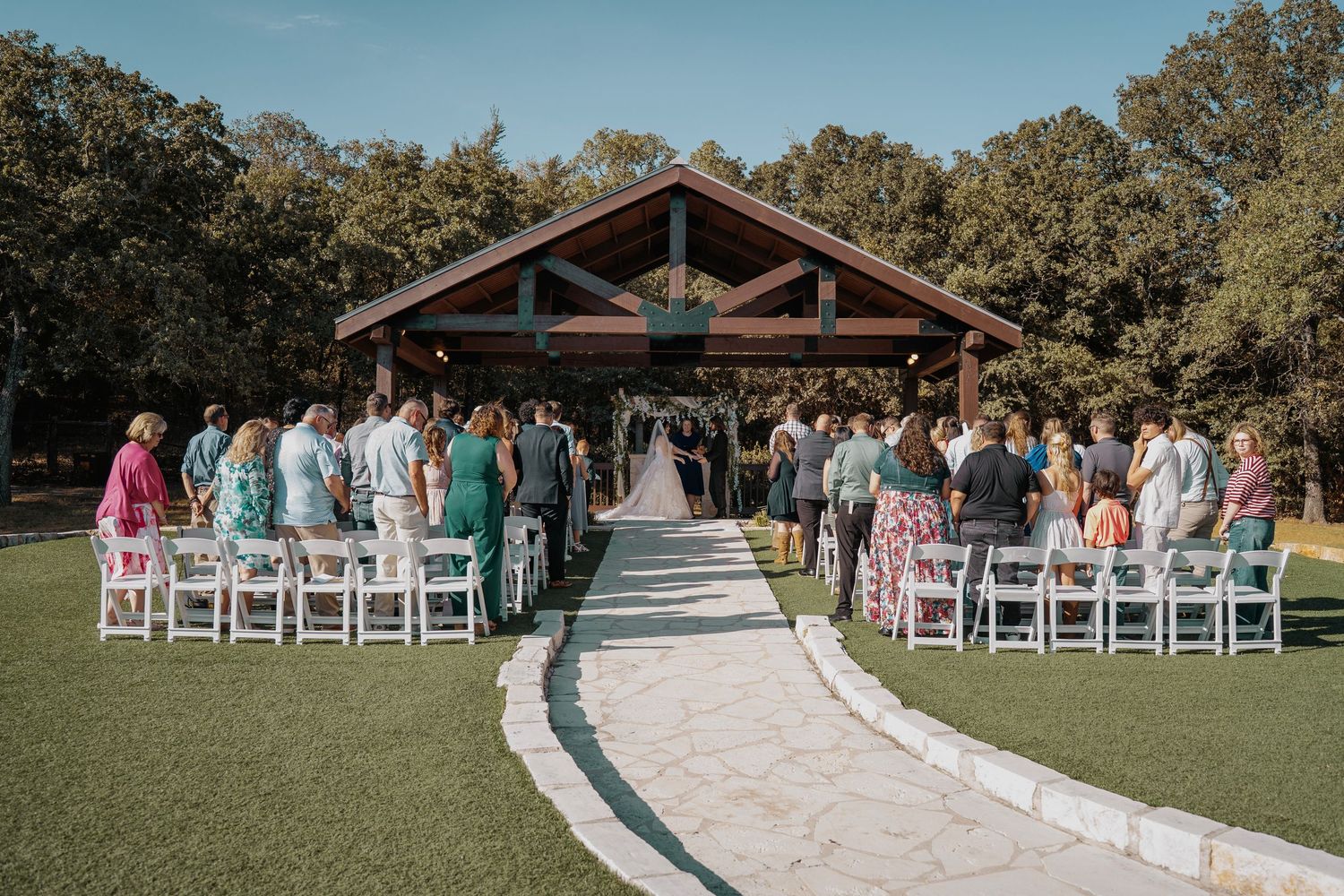 Outdoor wedding ceremony under a rustic pavilion surrounded by lush trees with guests seated in white chairs on a sunny day.