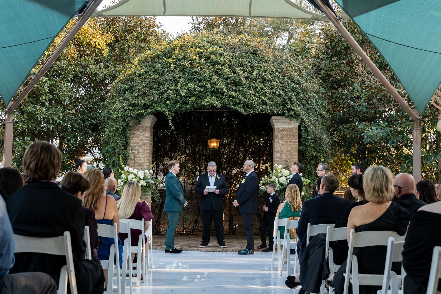 Outdoor wedding ceremony setup with white chairs and floral decorations under trees.