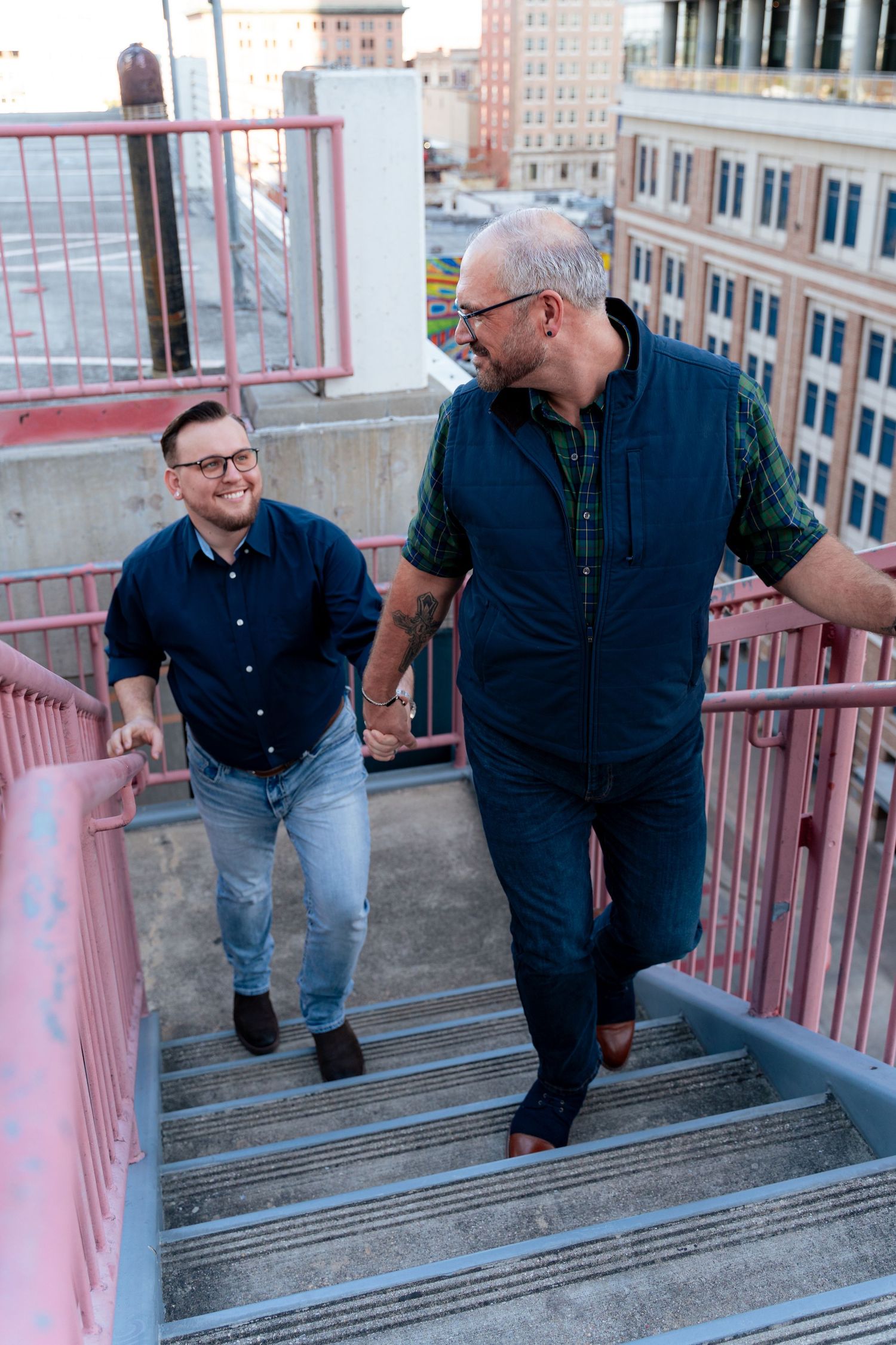 Two men walking up outdoor stairs in an urban setting, smiling at each other, wearing casual blue outfits.