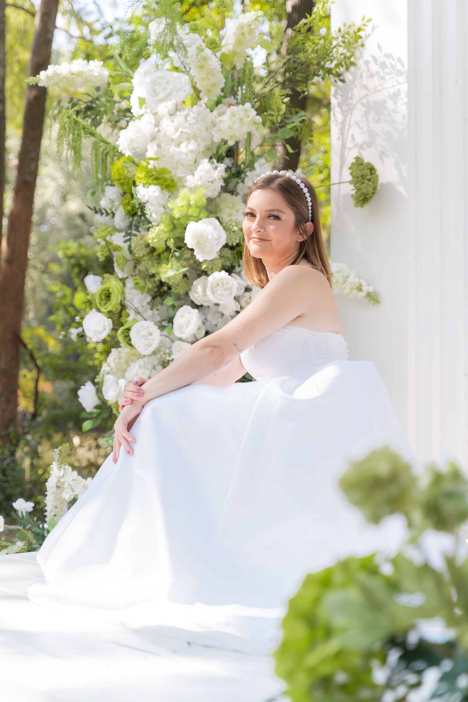 A bride in a white dress reclines on a white settee surrounded by elegant floral decorations.