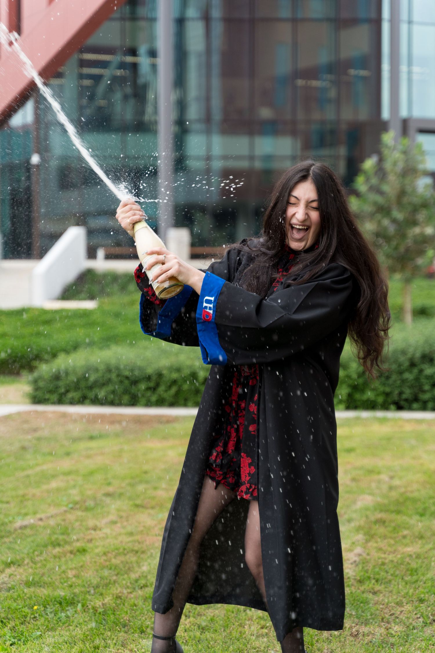 A joyful graduate in black academic gown sprays champagne outdoors, celebrating her graduation day.