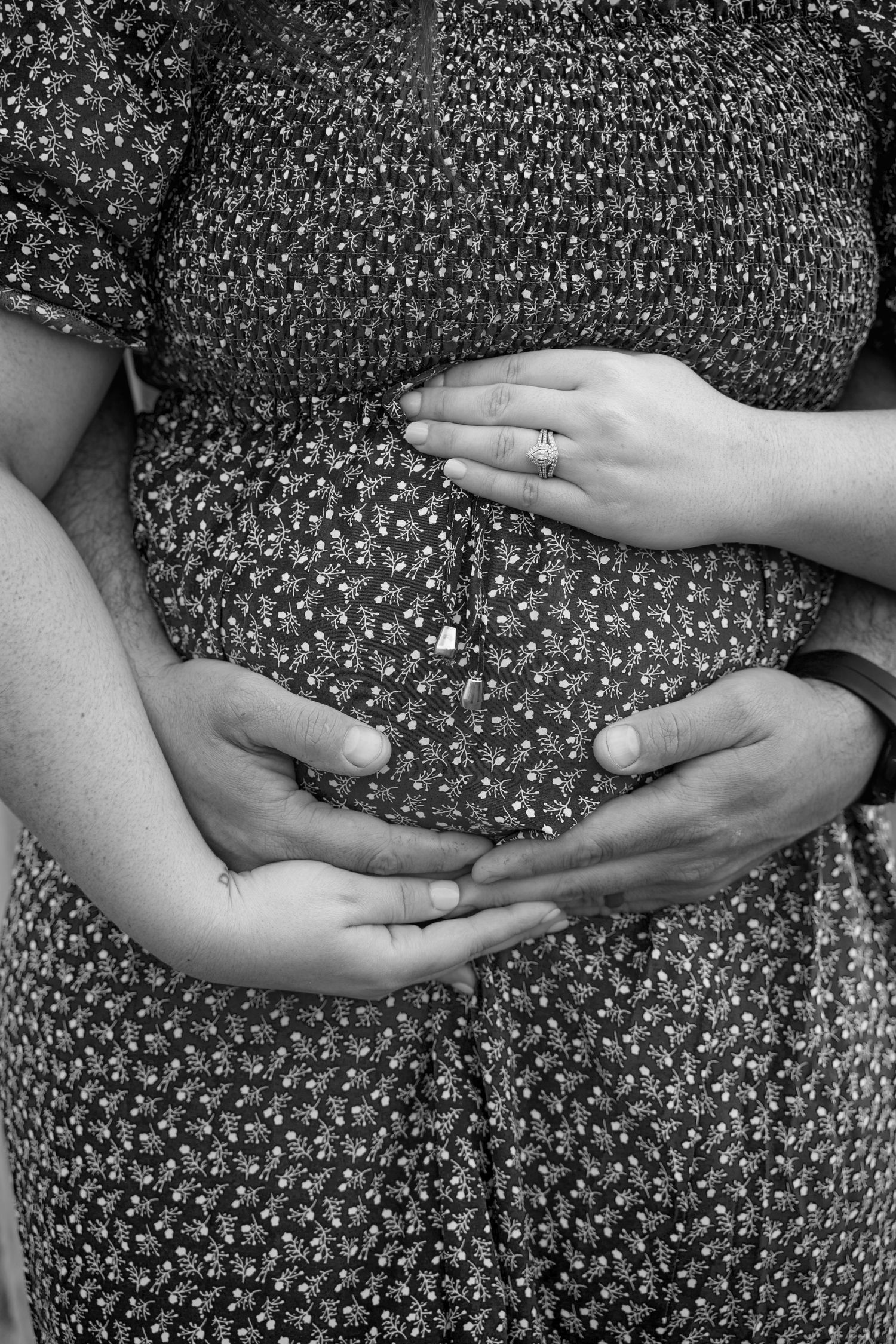 Black and white photo of a pregnant woman in floral dress with two pairs of hands cradling her baby bump.