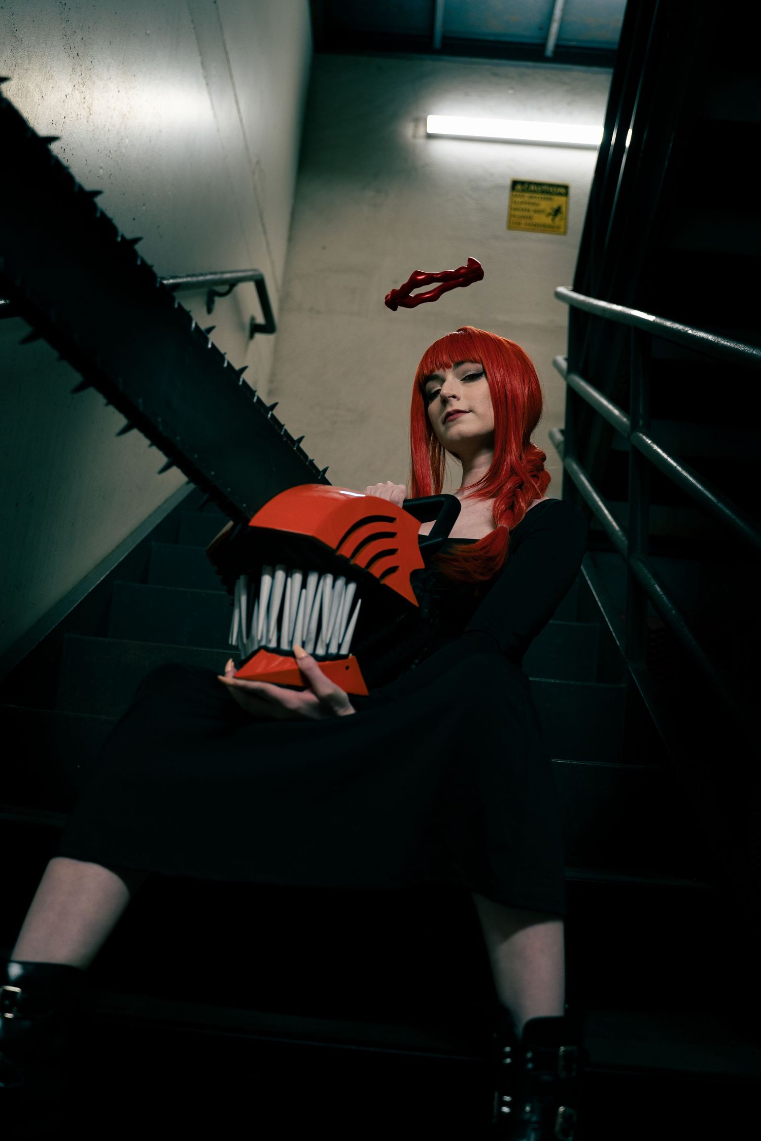 Person cosplaying as Chainsaw Man character Power, wearing red wig and holding chainsaw prop on dark staircase.