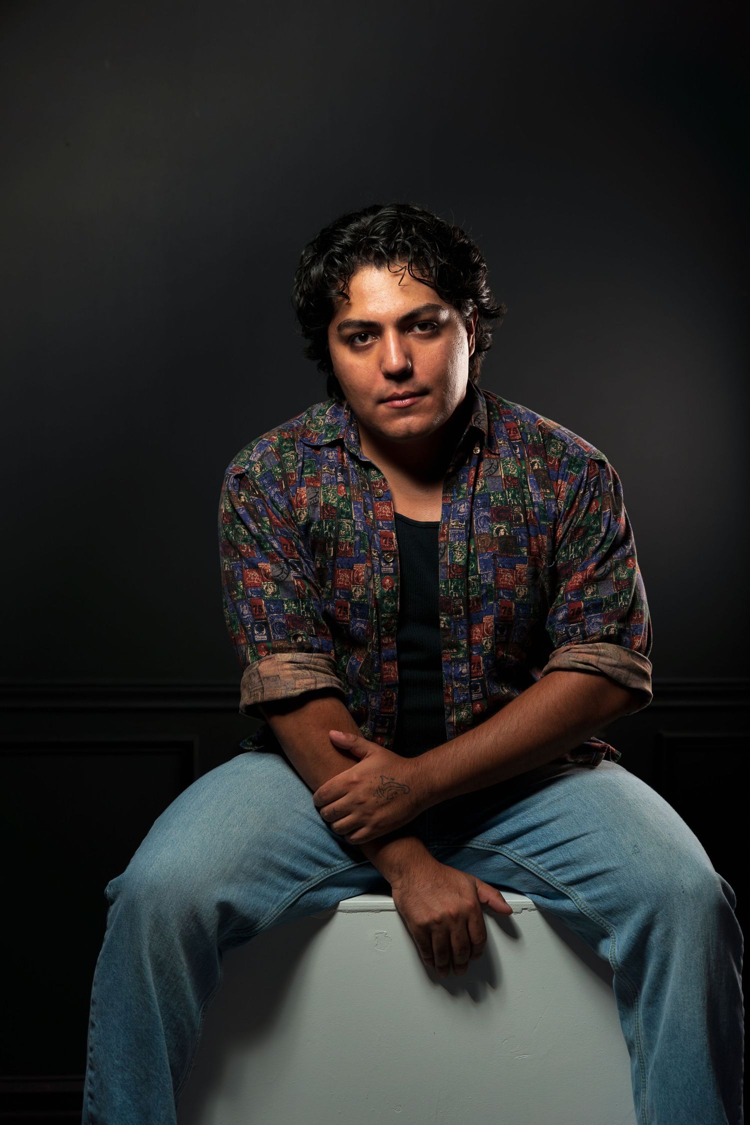 Young man with curly hair in colorful floral shirt sitting on white surface against dark background.