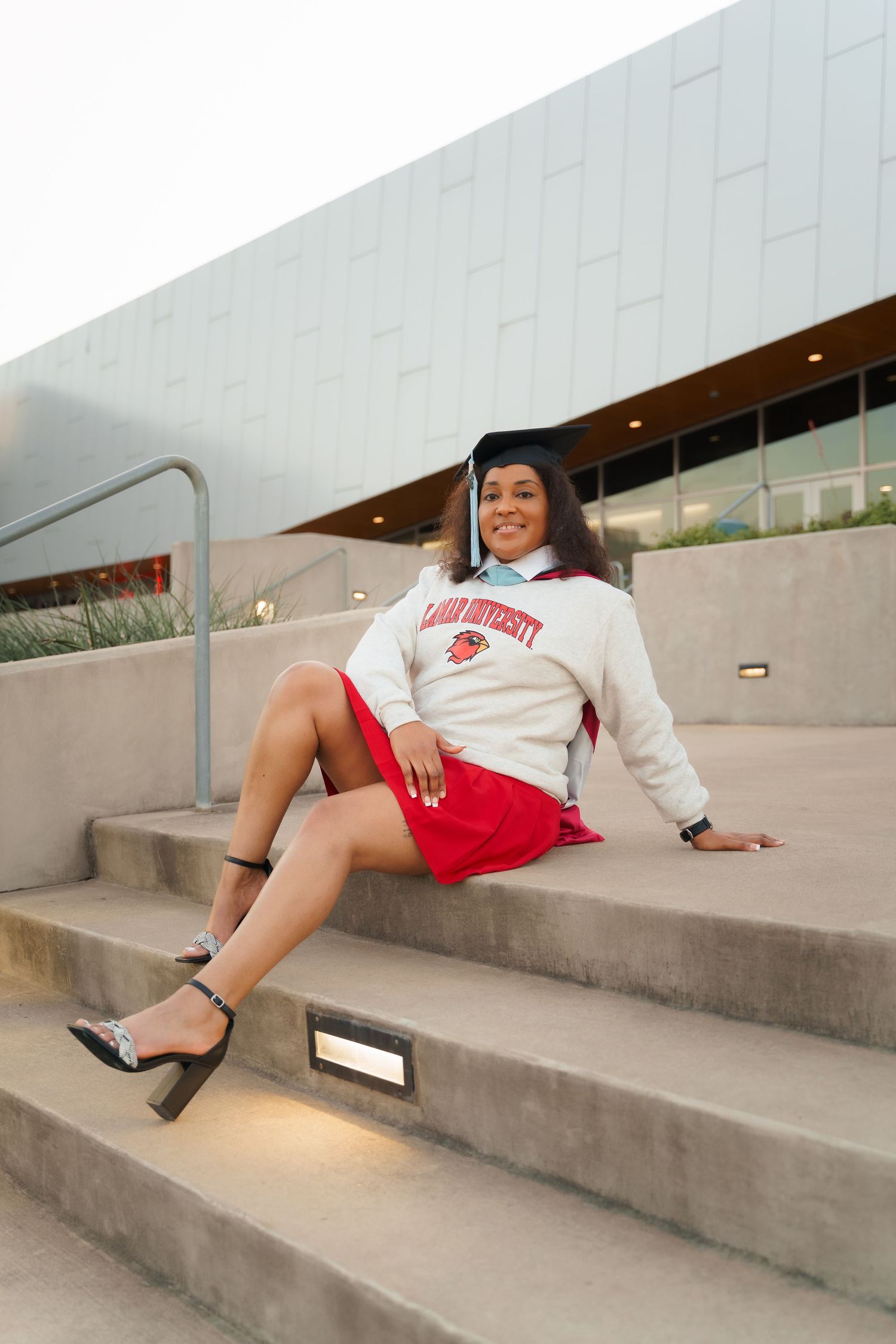 Young woman in graduation cap and university sweatshirt with red skirt sitting on concrete steps smiling.