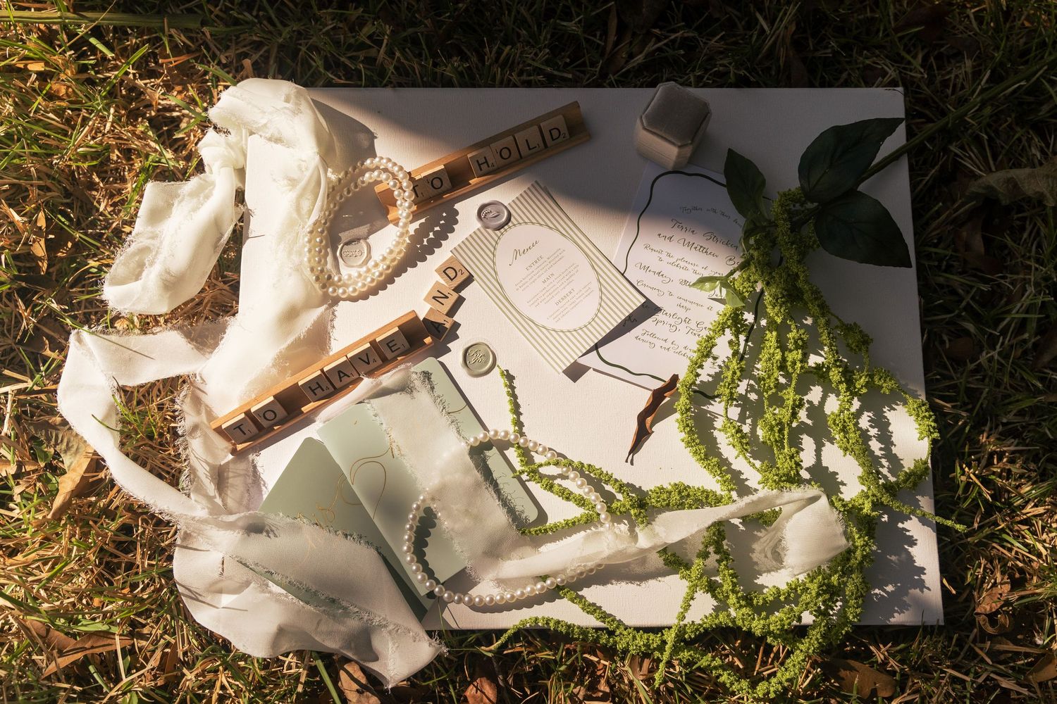 Flat lay of wedding accessories on grass, including pearls, a watch, greenery, photos, and elegant stationery.