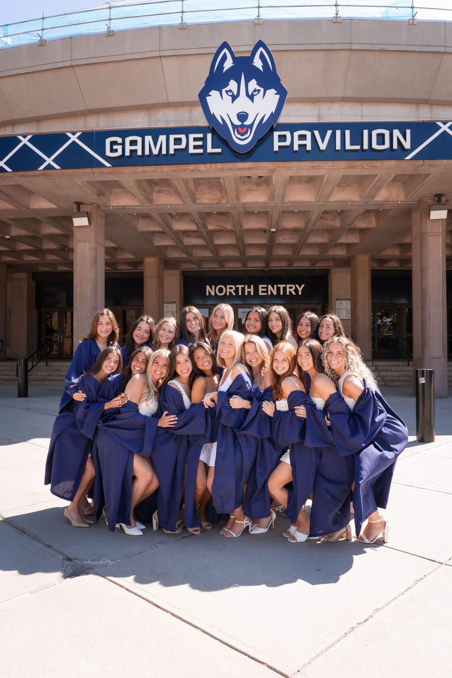 Group of female graduates in navy gowns posing outside Gampel Pavilion at UConn on graduation day.