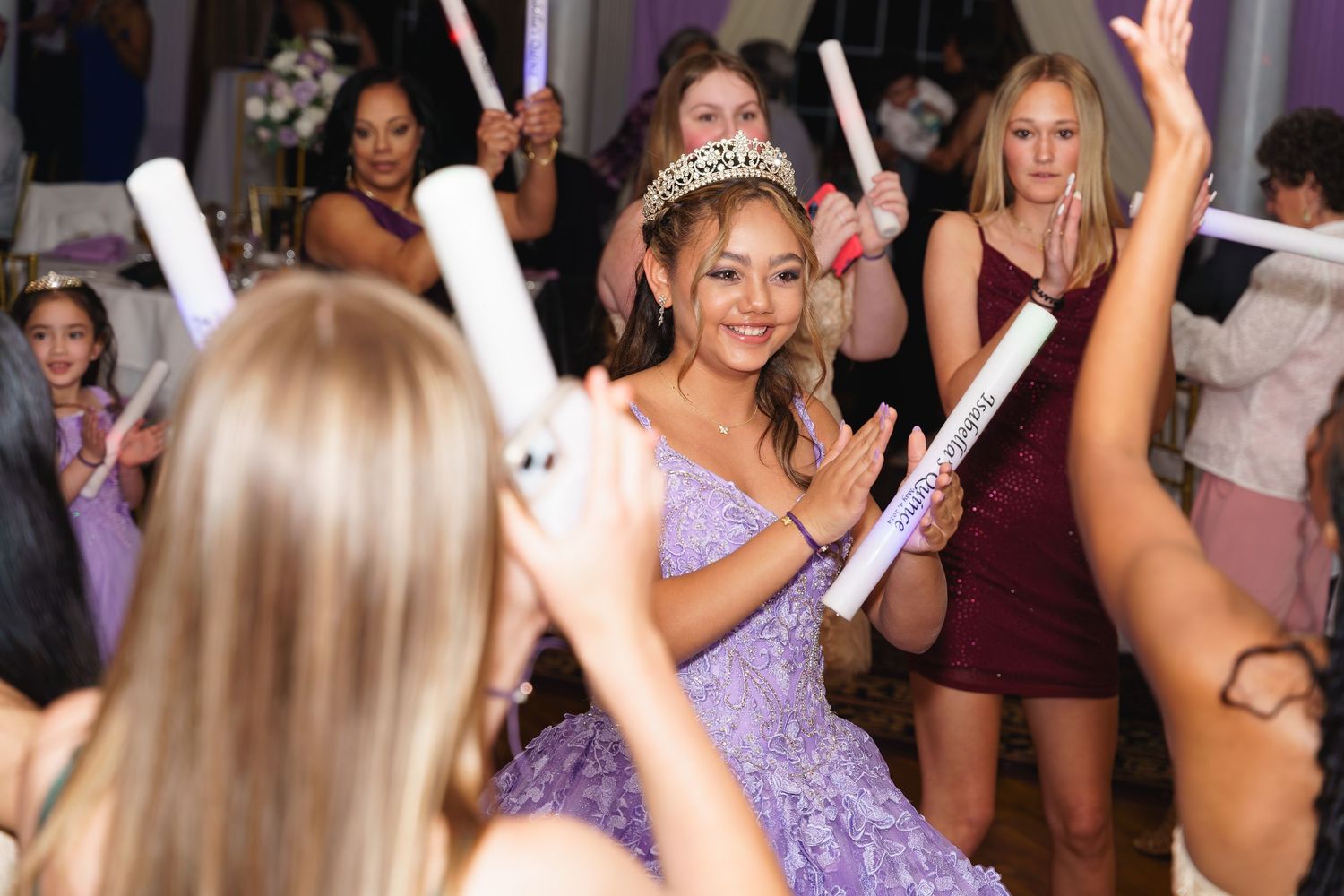 Young woman in lavender gown wearing tiara celebrates quinceañera with guests holding light-up wands at elegant party.