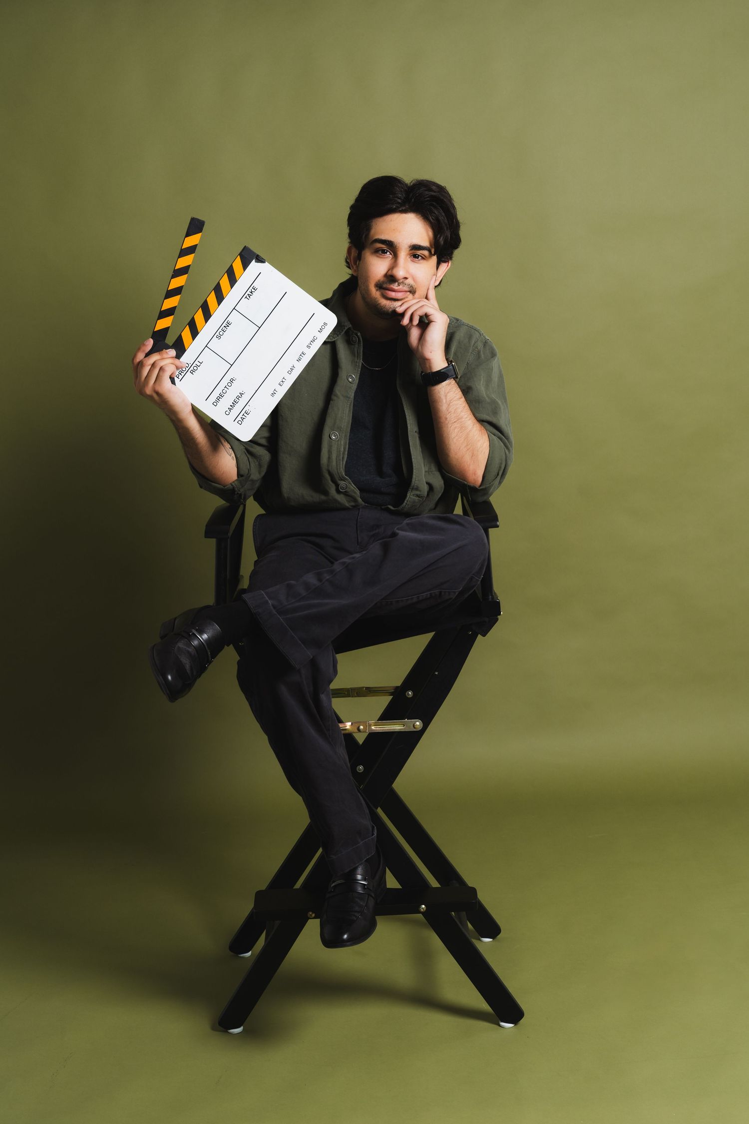 Young man in green shirt sits in director's chair holding film clapperboard on olive background.