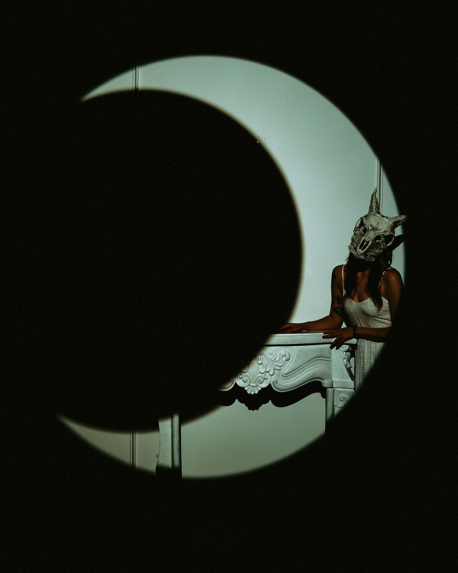 Person wearing animal skull mask posed behind a crescent moon shadow cutout in dramatic dark lighting.