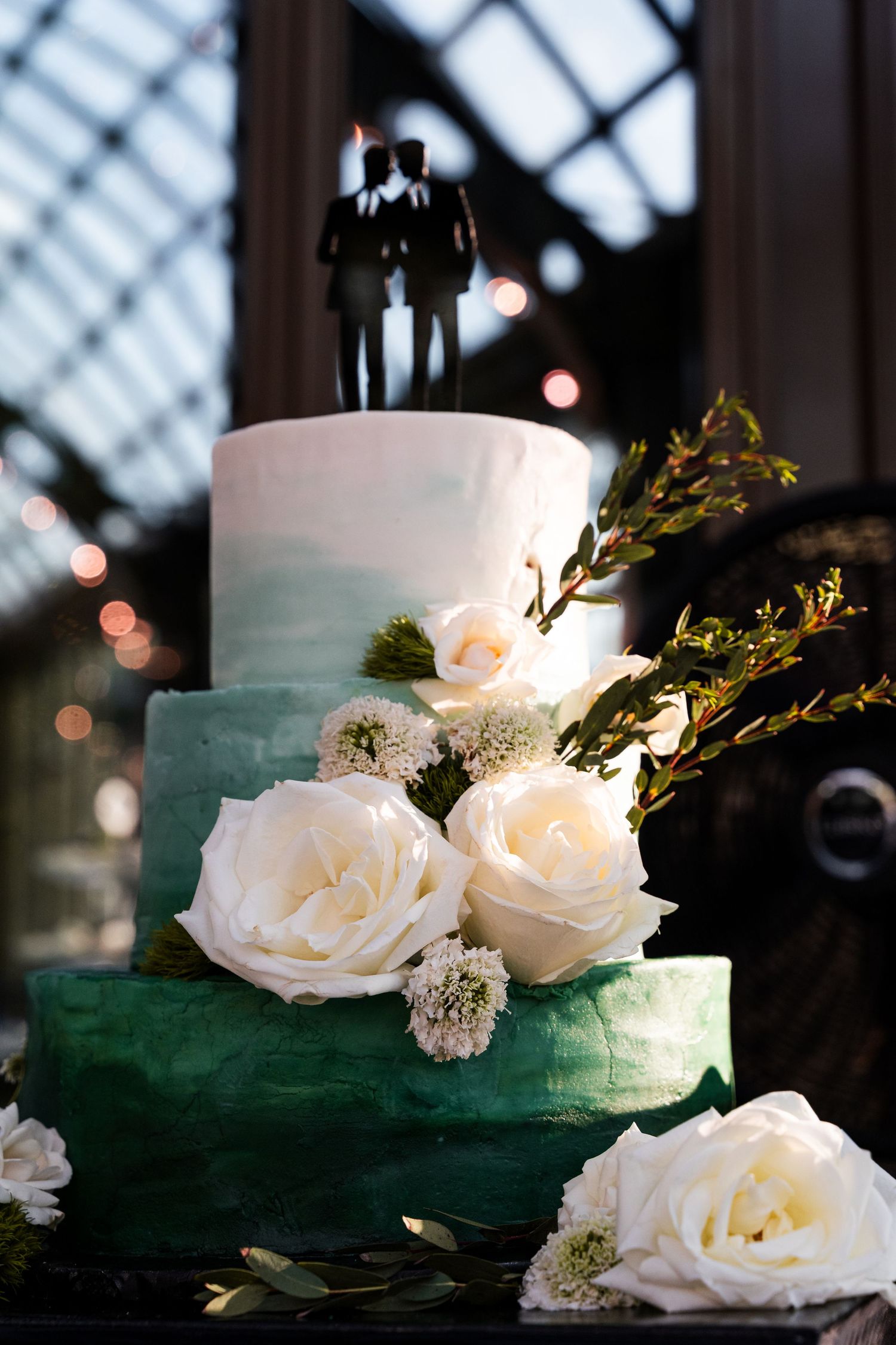Three-tier green ombre wedding cake with white roses and same-sex couple silhouette topper.