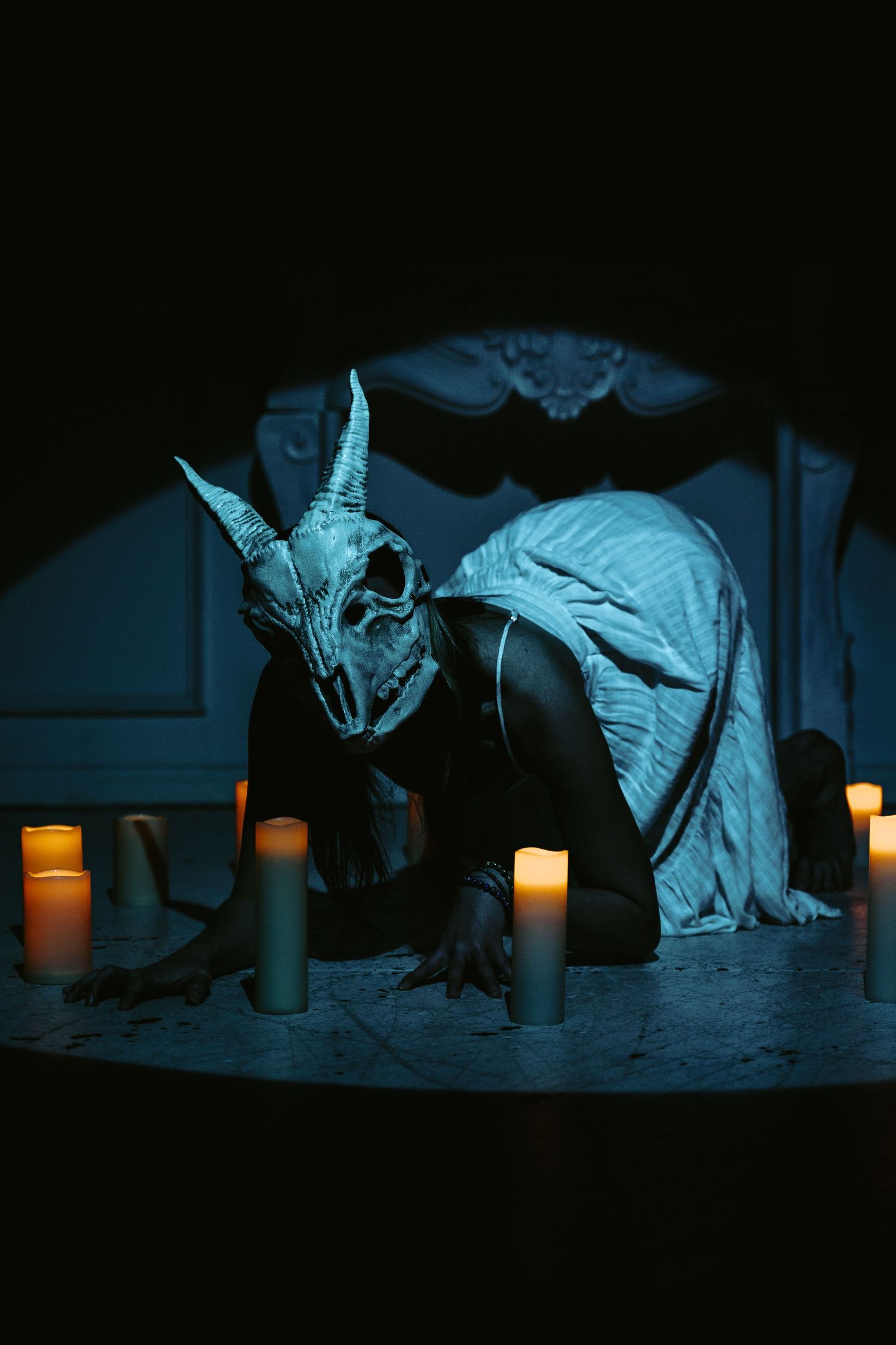 Person wearing a dragon skull mask crouching among lit candles in a dark, moody gothic setting with blue lighting.
