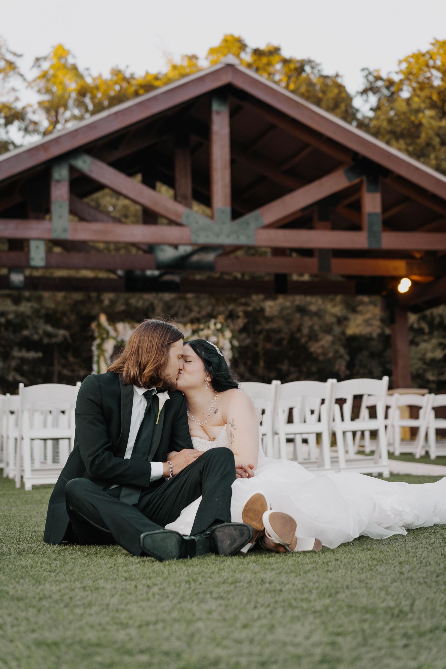 Bride and groom sitting on grass kissing in front of white chairs and rustic wooden pavilion at outdoor wedding venue.