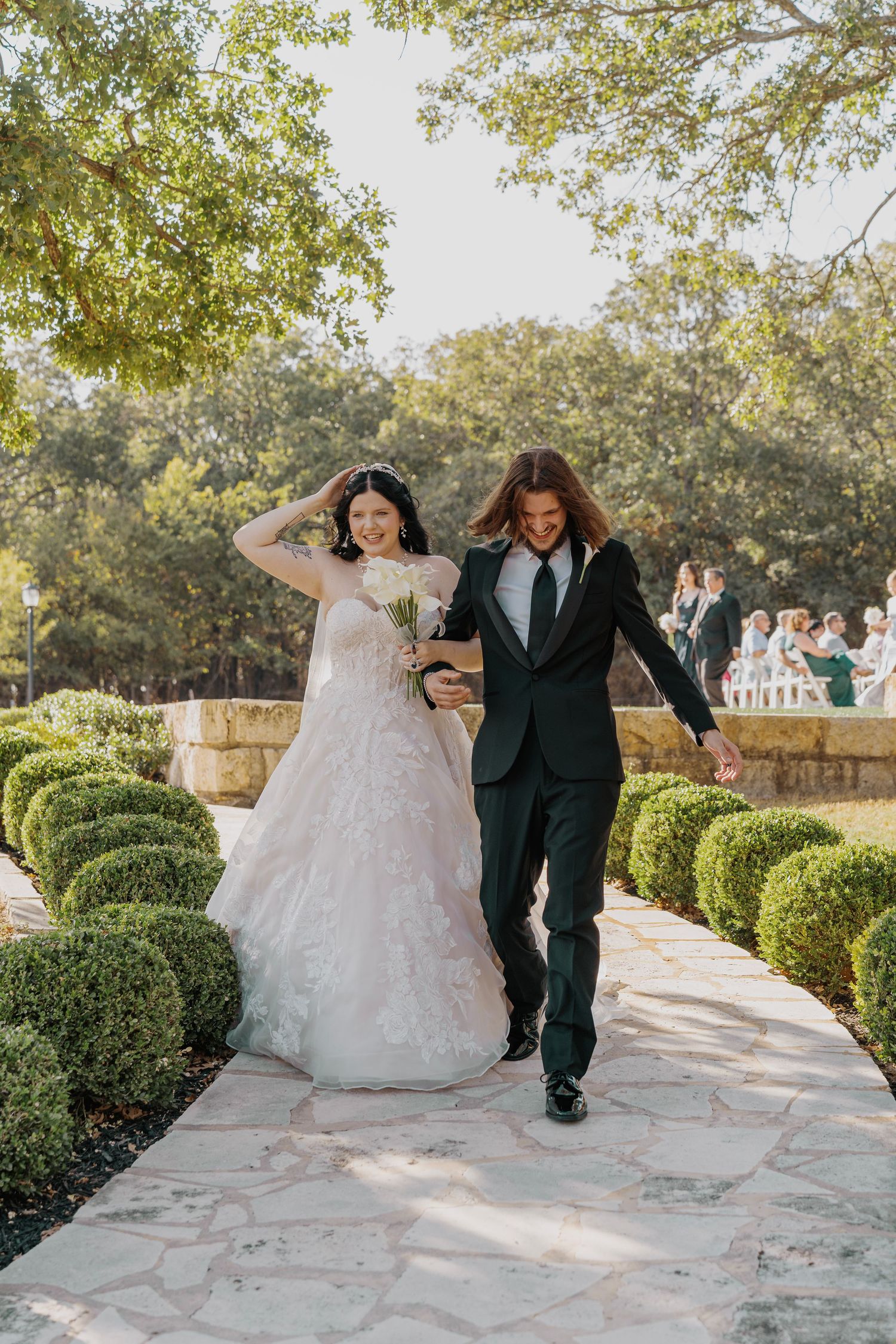 Bride in floral gown and groom in black tuxedo walk joyfully down garden path after wedding ceremony.