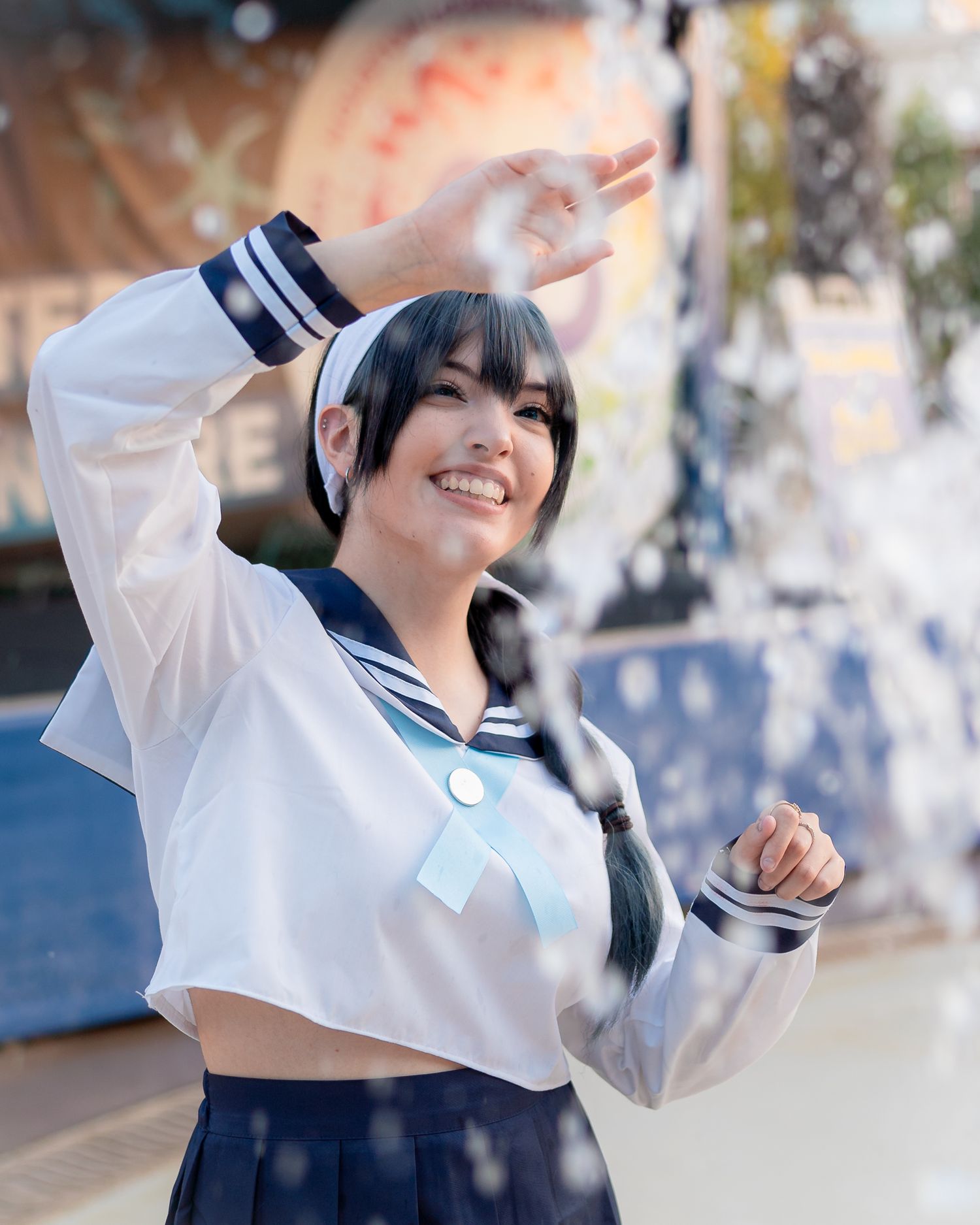 A smiling woman in a white sailor school uniform cosplay with navy skirt poses outdoors at an anime event.