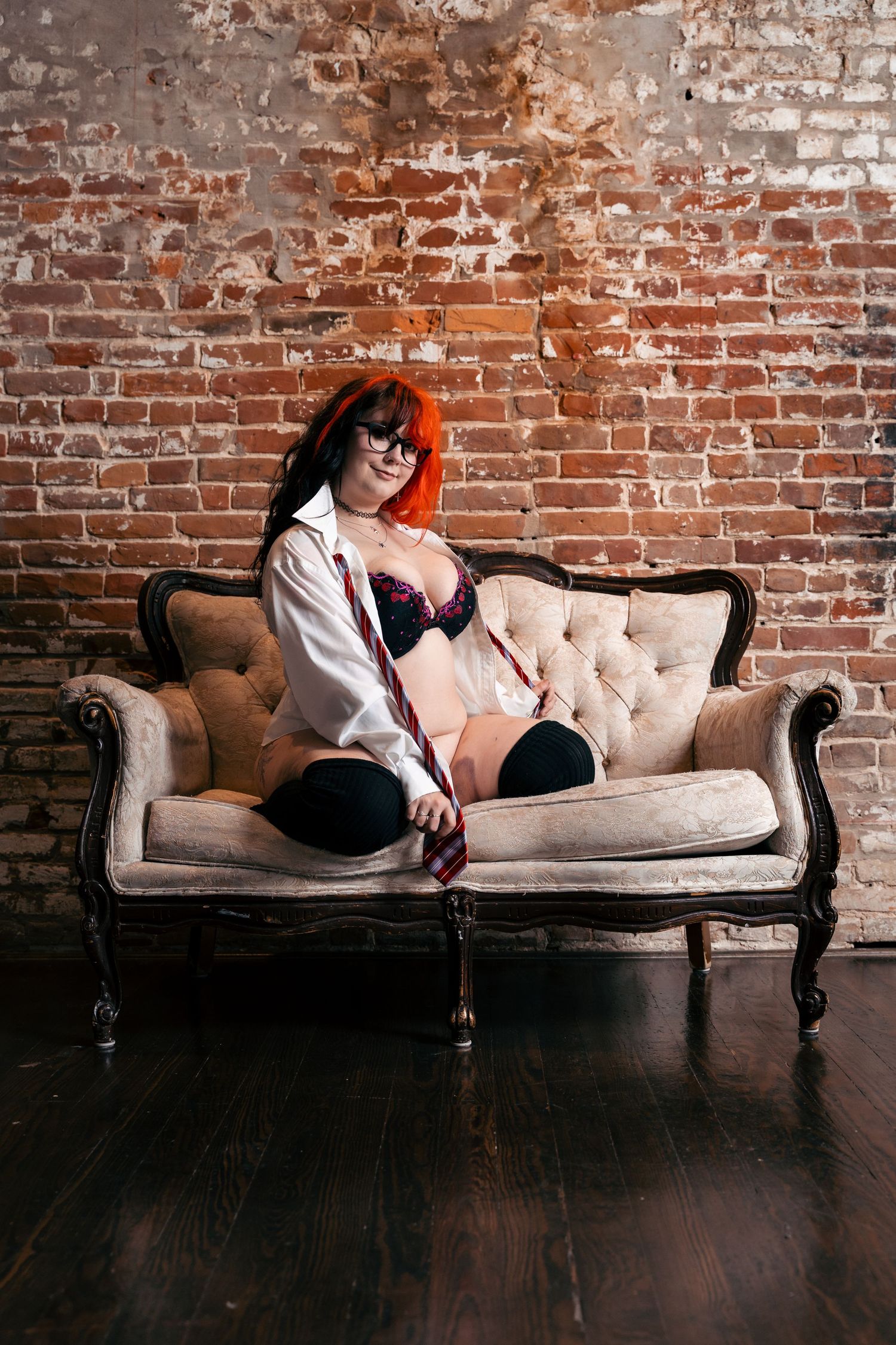 Woman with red hair and glasses in lingerie sitting on vintage sofa against brick wall.