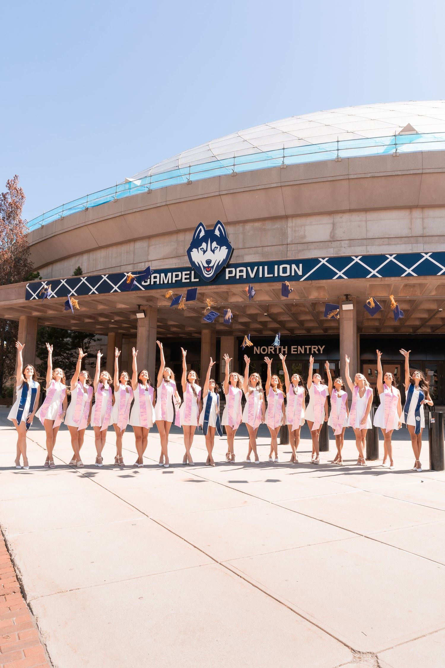UConn women's team in pink dresses posing outside Gampel Pavilion with arms raised on a sunny day.