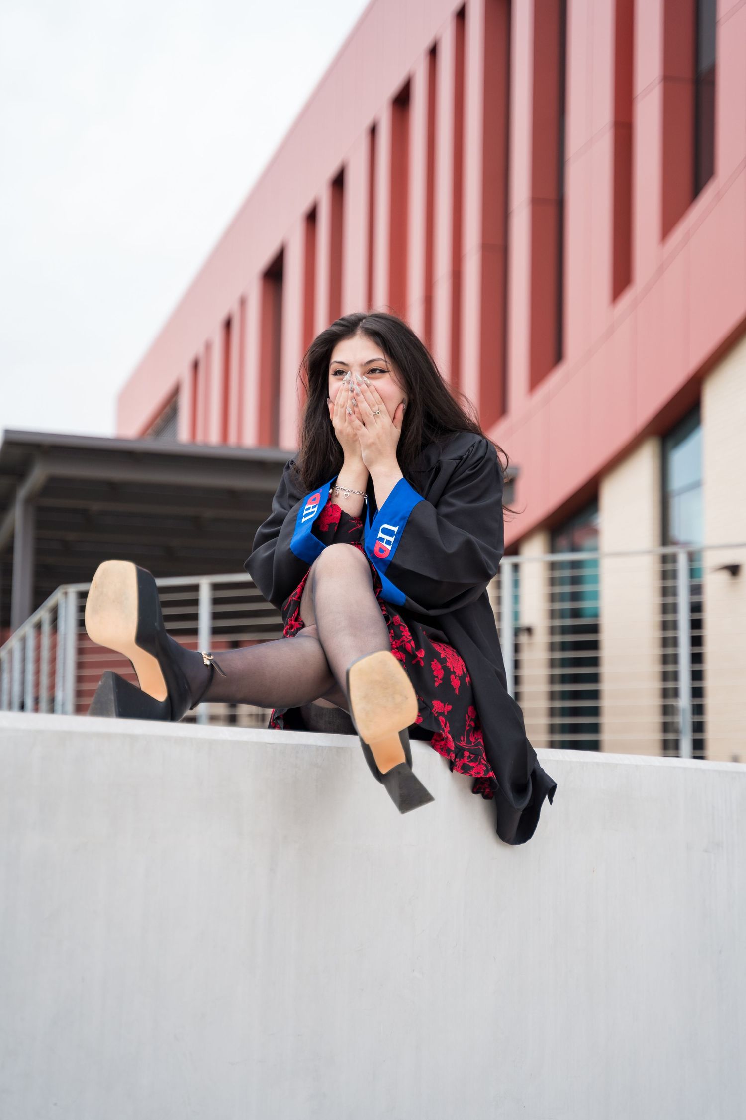 A young woman in graduation gown and blue sash sits on a wall, covering her mouth in excitement.