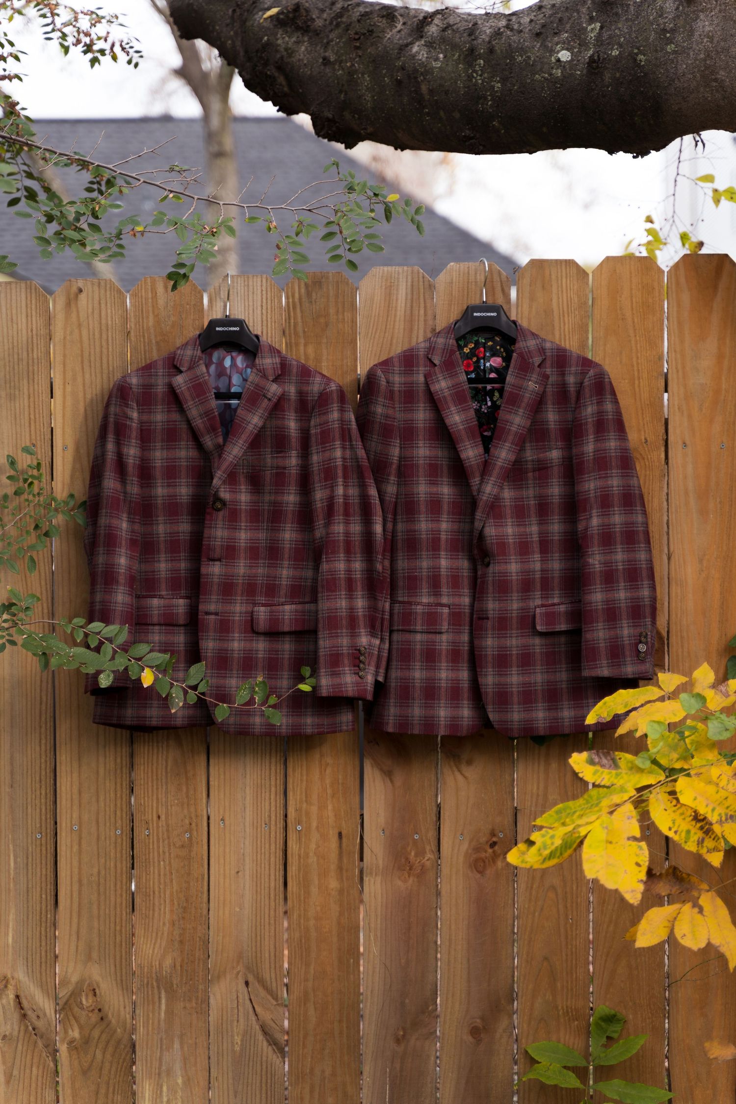 Two plaid brown sport coats hanging on wooden fence with autumn leaves in background.