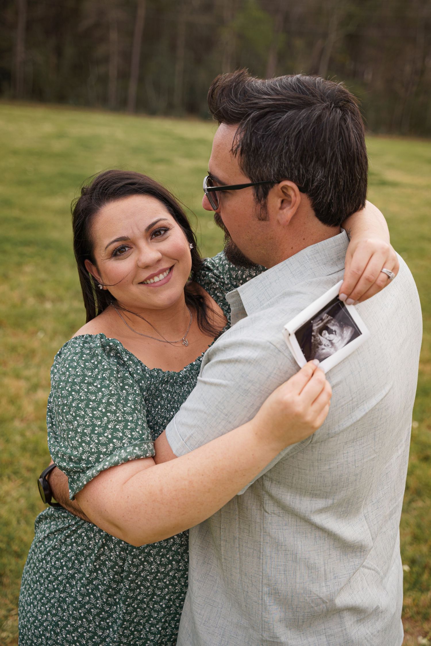 Happy couple embracing outdoors holding ultrasound photo, pregnancy announcement in green field setting.