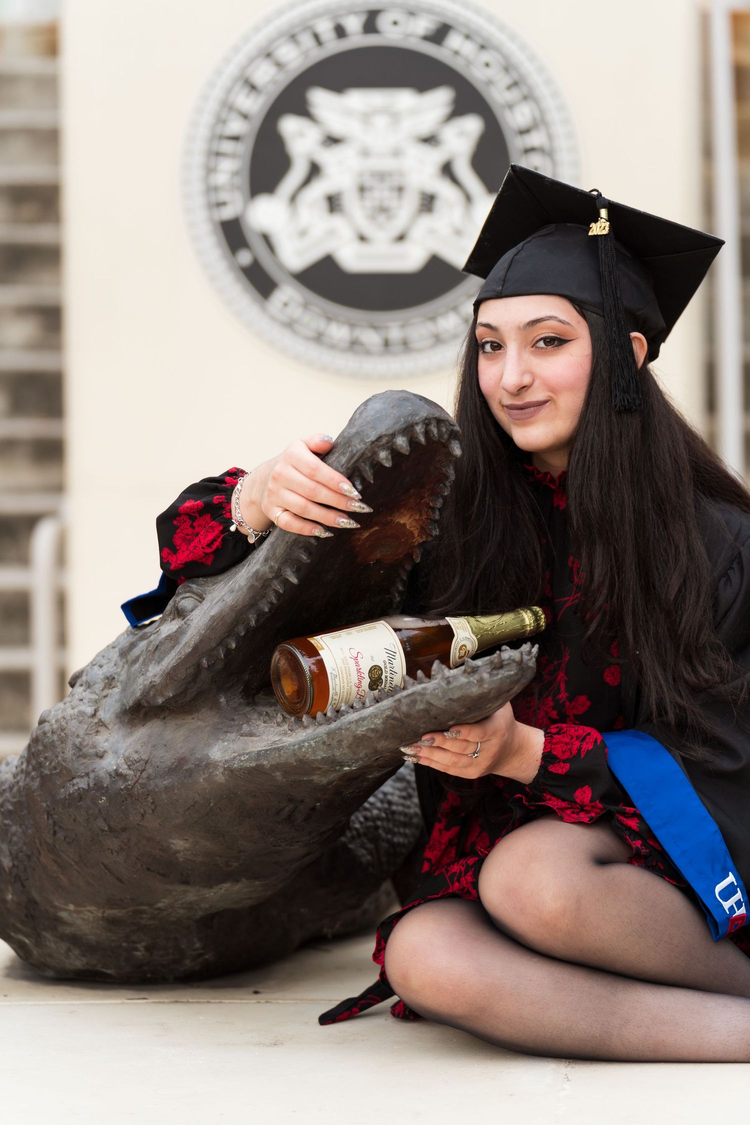 Graduate in cap and gown poses with alligator statue holding bottle at University of Houston.