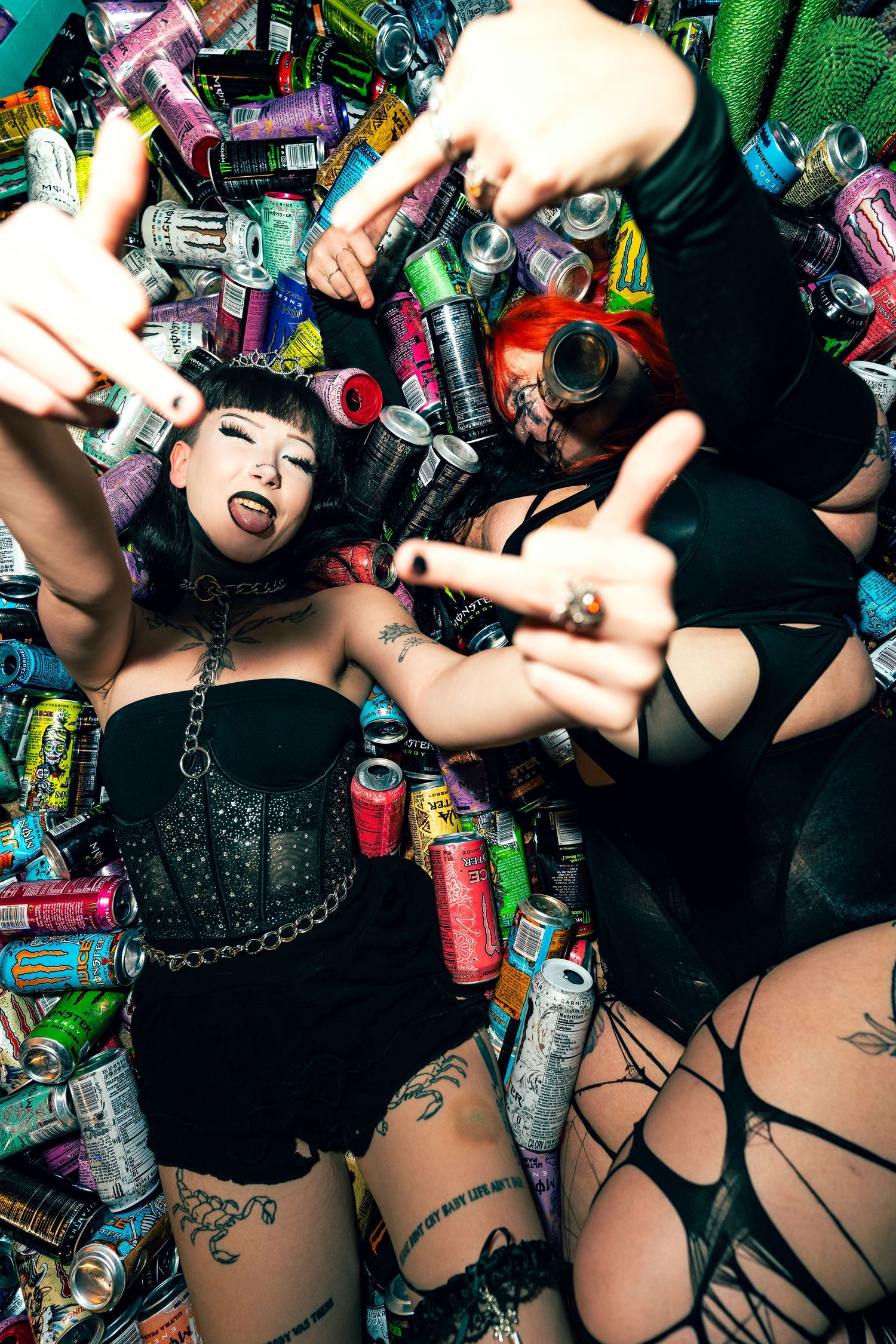 Two alt women in black outfits pose among colorful energy drink cans, making rock gestures with tattoos visible.