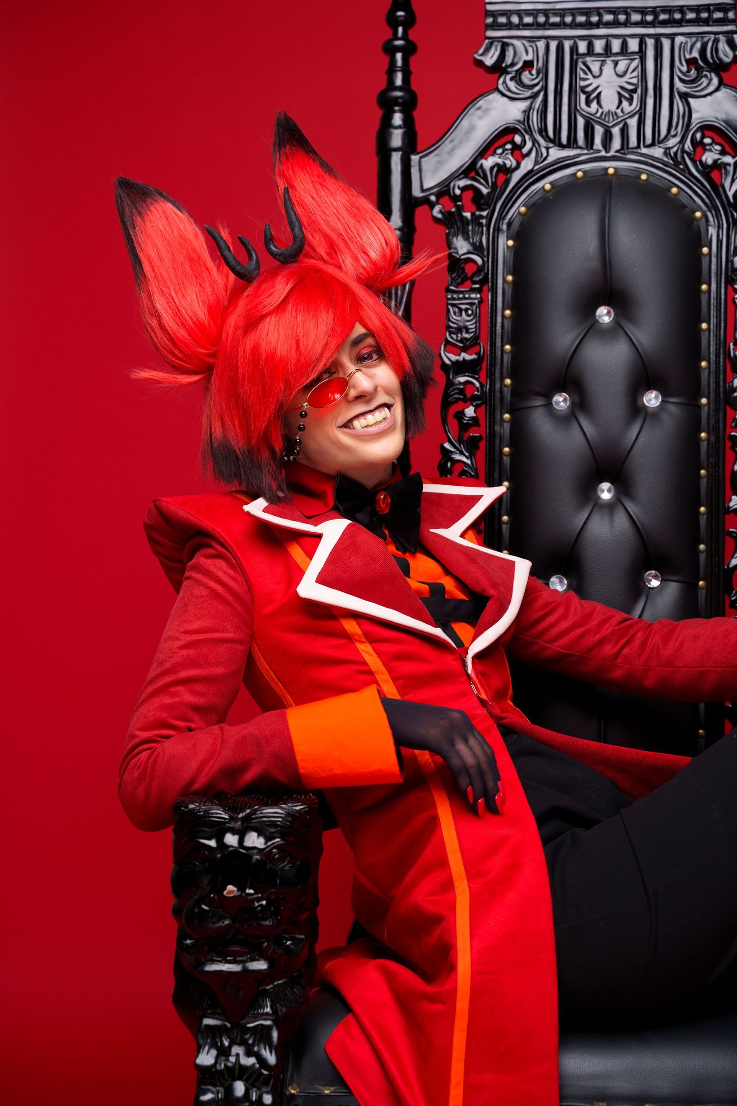 Person in red fox cosplay costume with elaborate wig poses beside ornate black throne against red background.