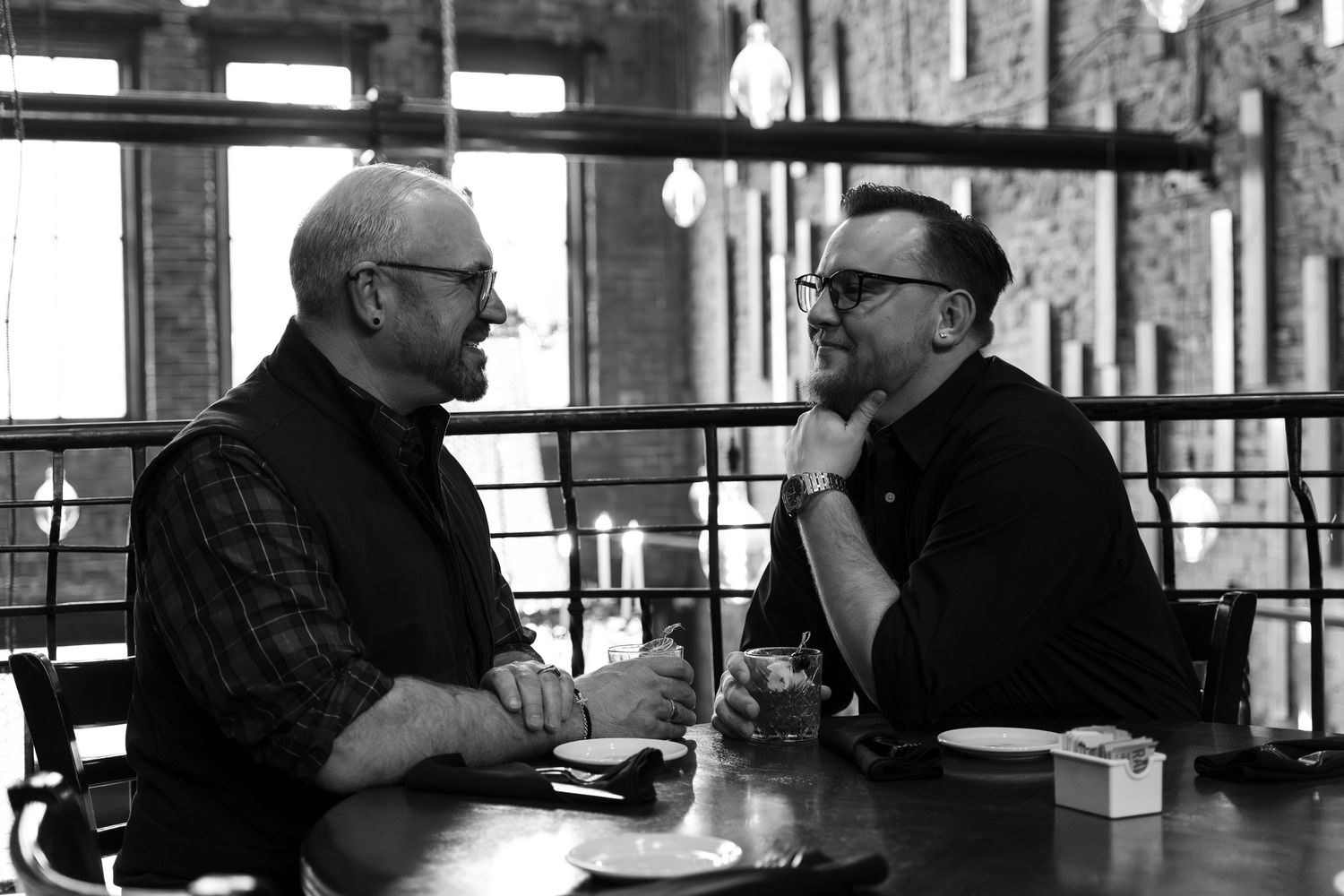 Two men in glasses share a conversation at a restaurant table in a black and white photo.