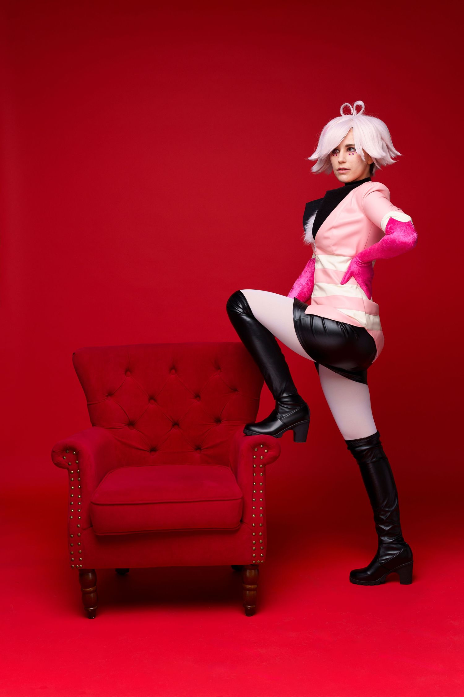 Woman in pink and black cosplay costume with white wig and knee-high boots posing beside a red tufted armchair.