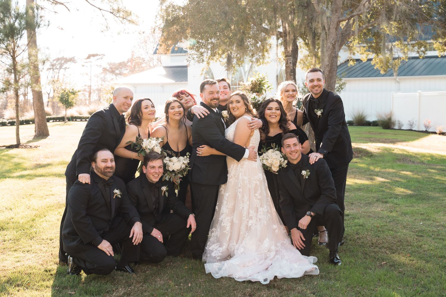 Wedding party in black attire poses playfully on grass for group photos.