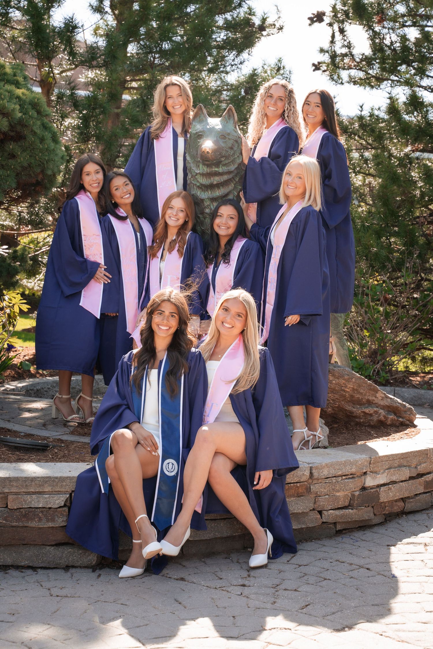 Group of female graduates in navy blue robes and pink stoles posing near a wolf statue outdoors.