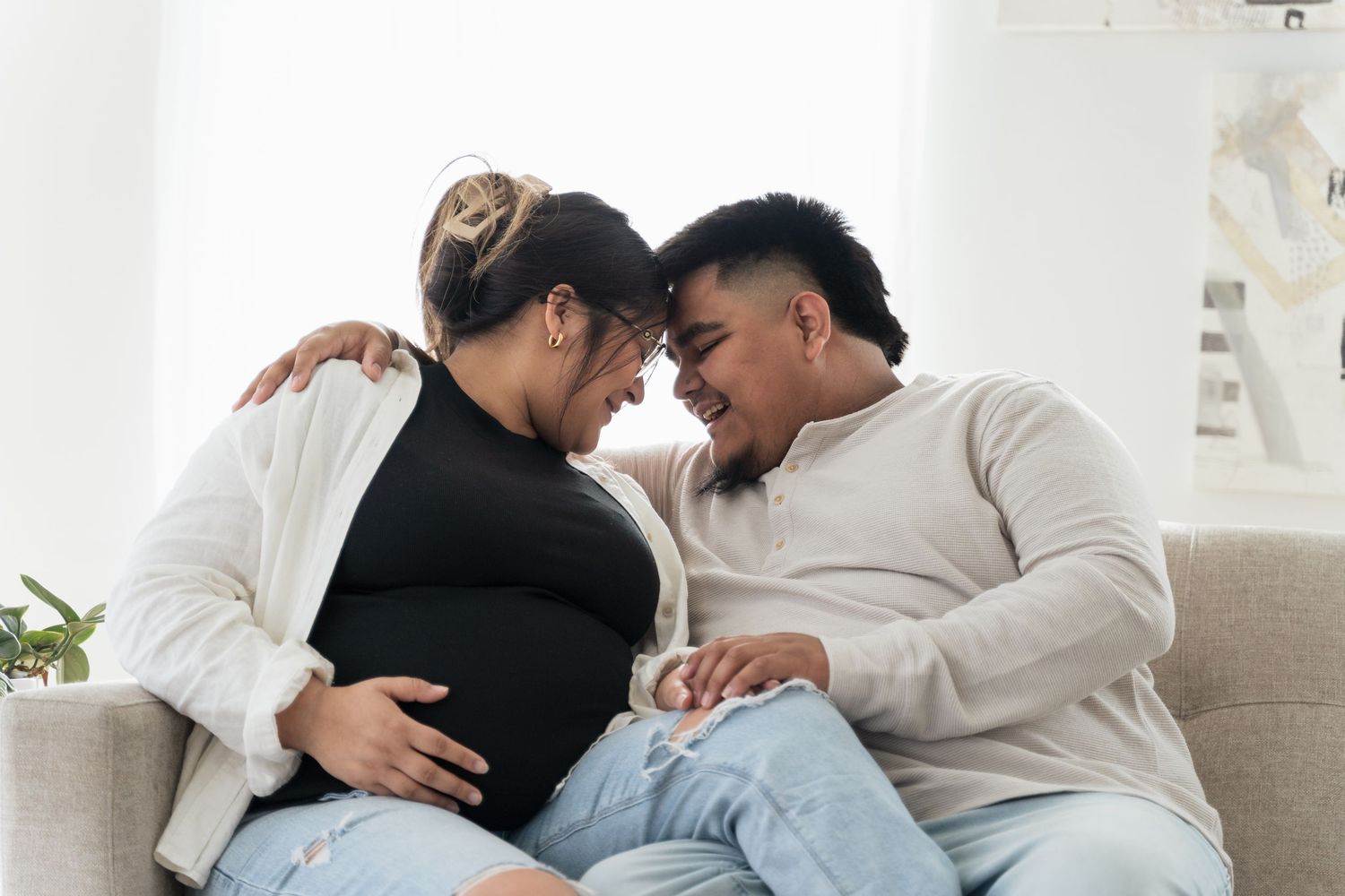 Happy pregnant couple sitting on sofa, touching baby bump and smiling at each other in bright living room.