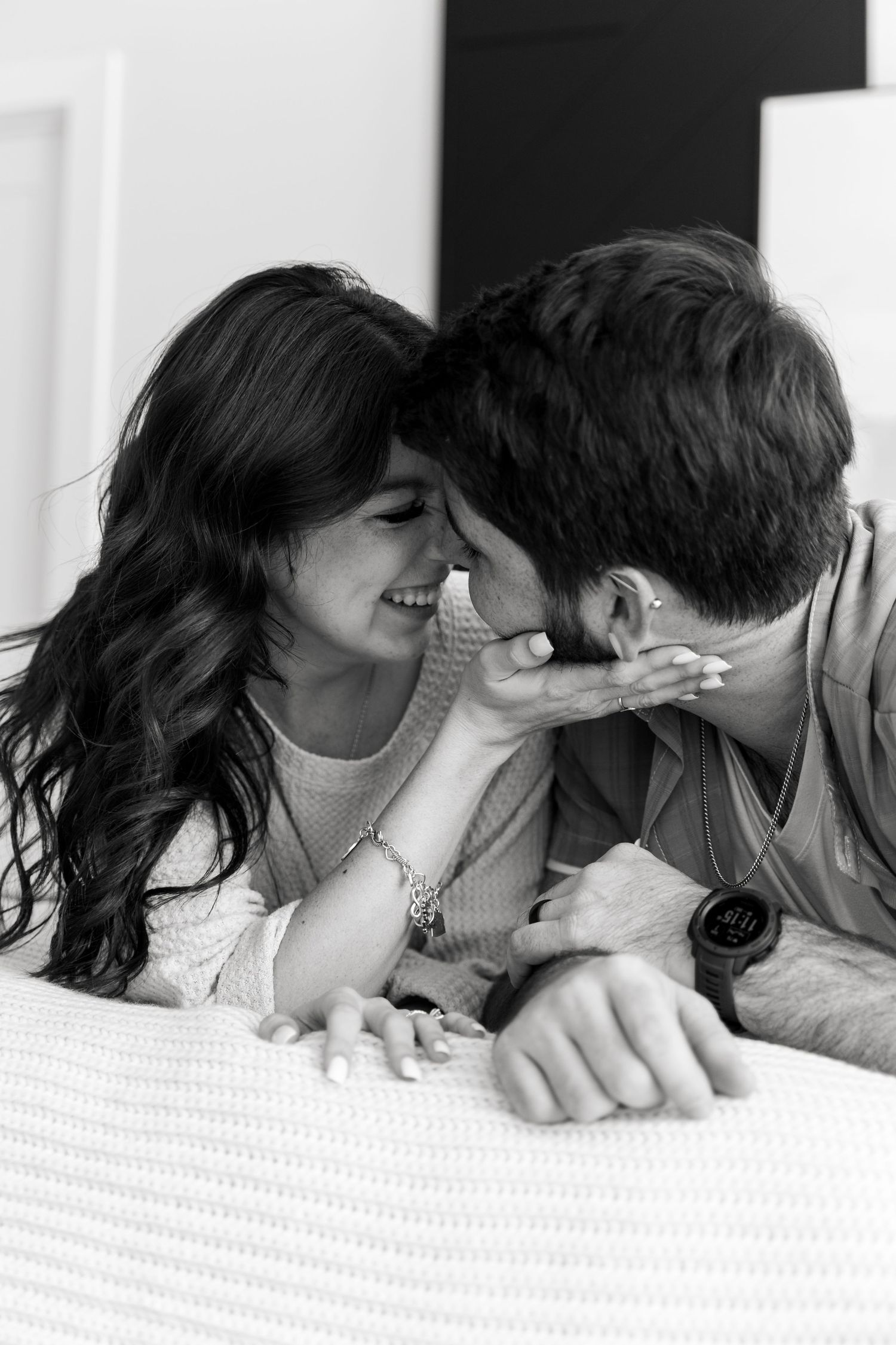 Black and white photo of a smiling couple sharing a tender moment lying on a bed, woman touching mans face.