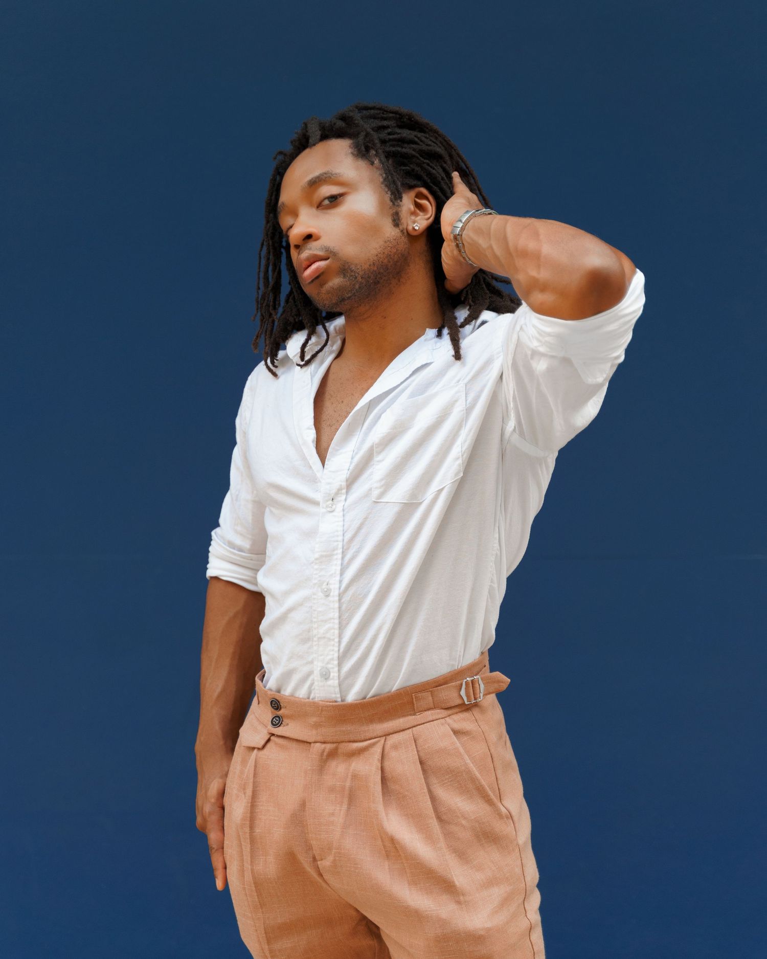 Young man with locs wearing a white button-up shirt and tan trousers posing against a blue background.