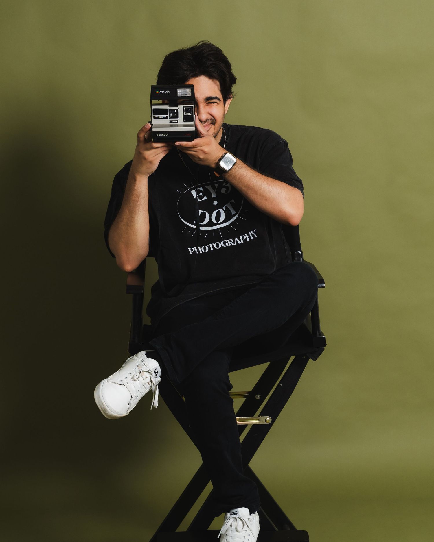 A young man in black outfit sits on a director's chair holding a Polaroid camera up to his face in a studio.