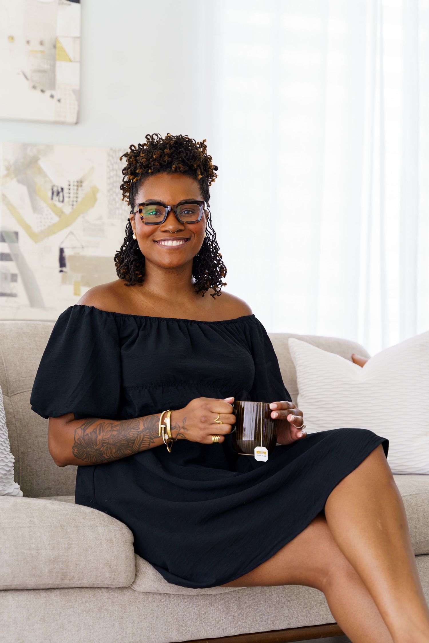 Smiling woman in black off-shoulder dress sitting on sofa holding a mug, wearing glasses and bracelet.