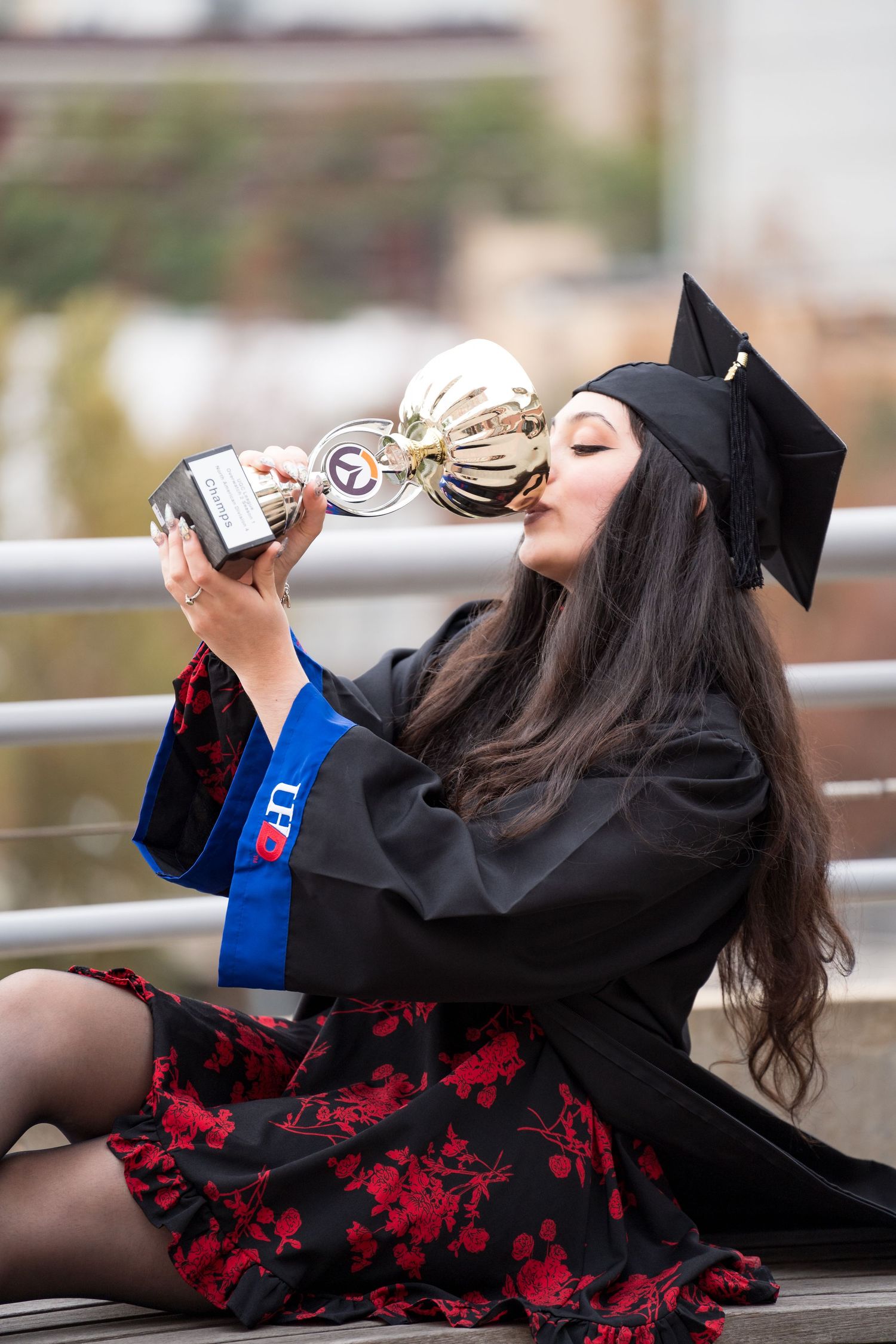 A young woman in graduation cap and gown kisses a trophy while sitting outdoors, celebrating her academic achievement.