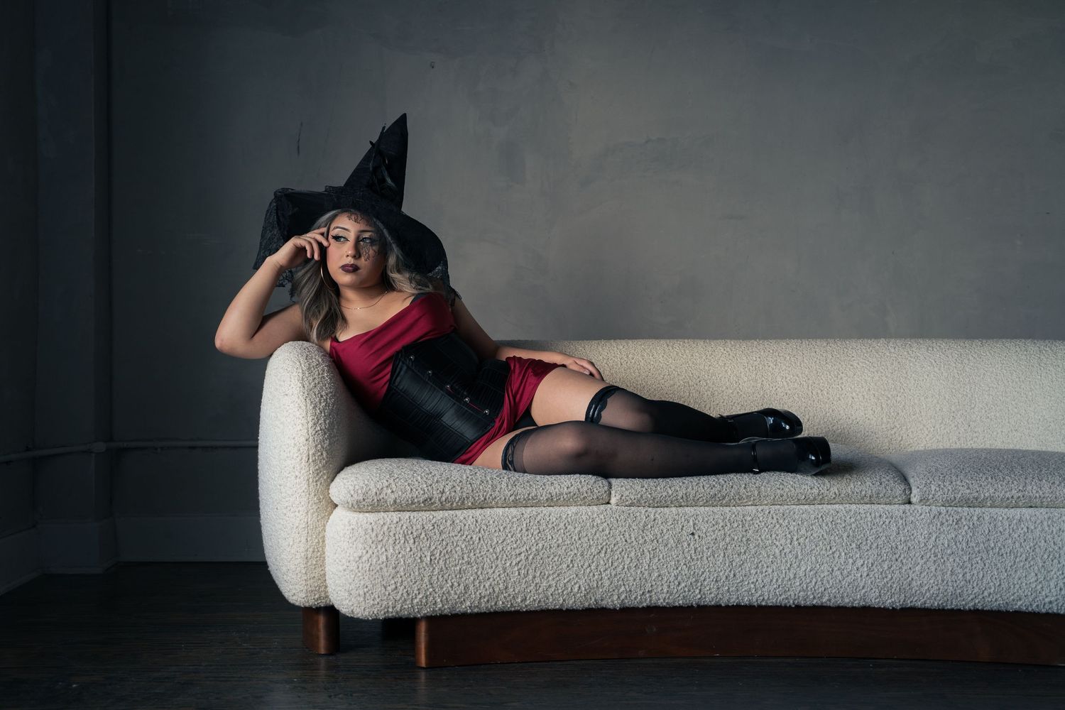 Woman in red corset, black stockings, and witch hat poses on white sofa for Halloween costume photo.