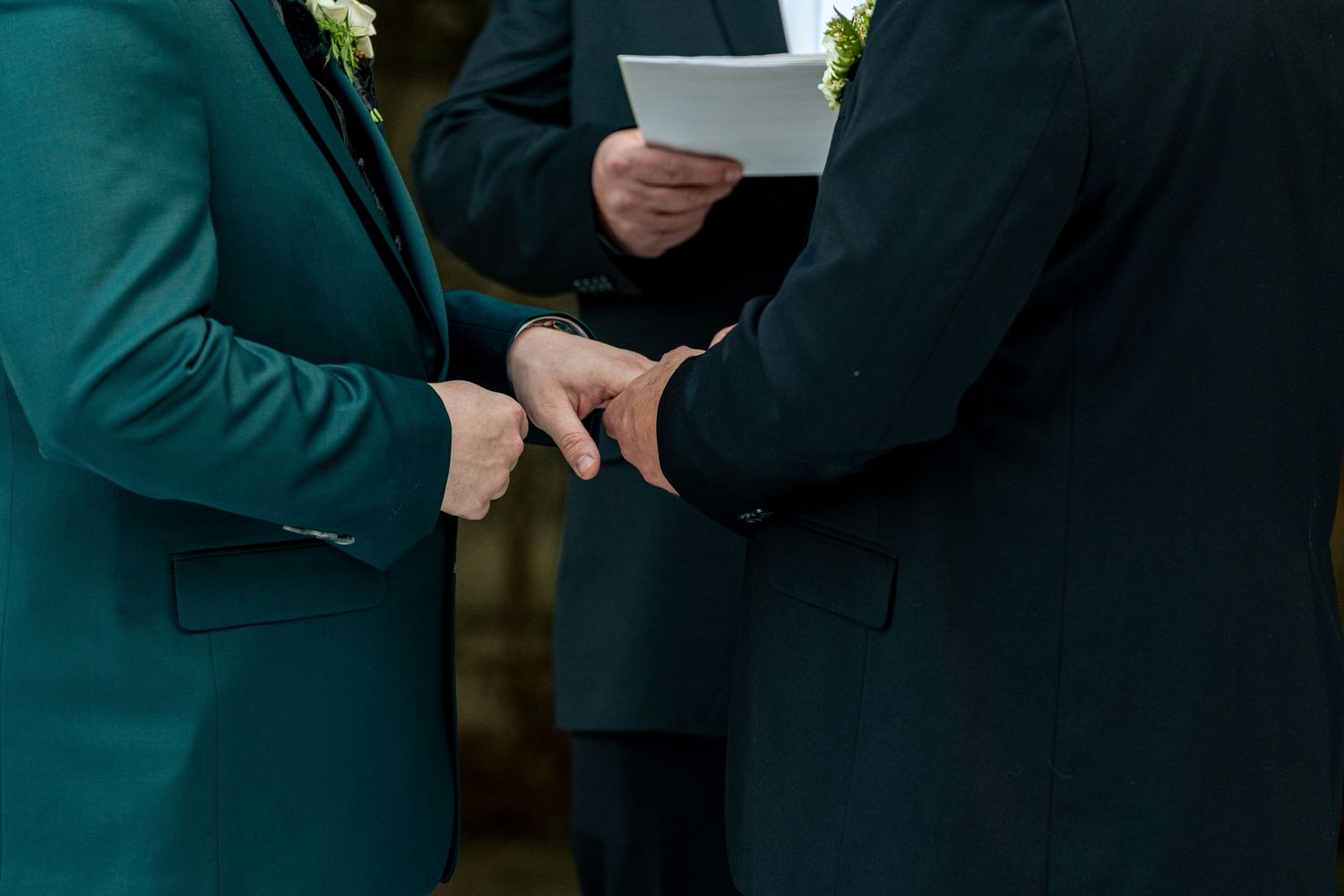Two grooms in dark suits exchange rings during a wedding ceremony officiated by a person holding vows.