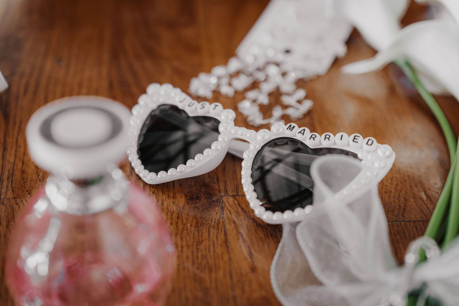 Heart-shaped pearl-trimmed bridal sunglasses with MARRIED charm on wooden table with perfume bottle and veil.