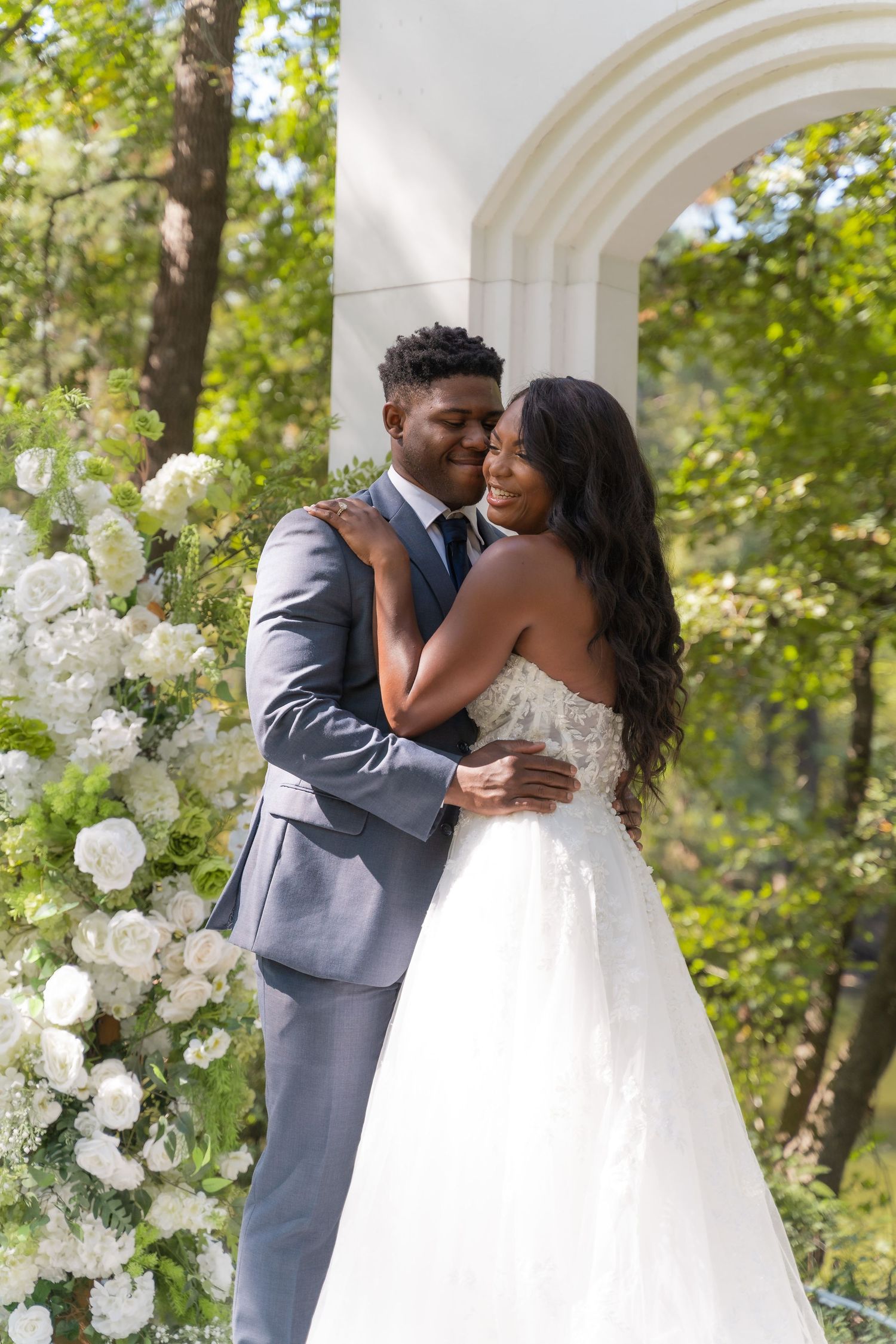 A Black bride and groom embrace outdoors near white floral arrangements and arched columns surrounded by lush greenery.