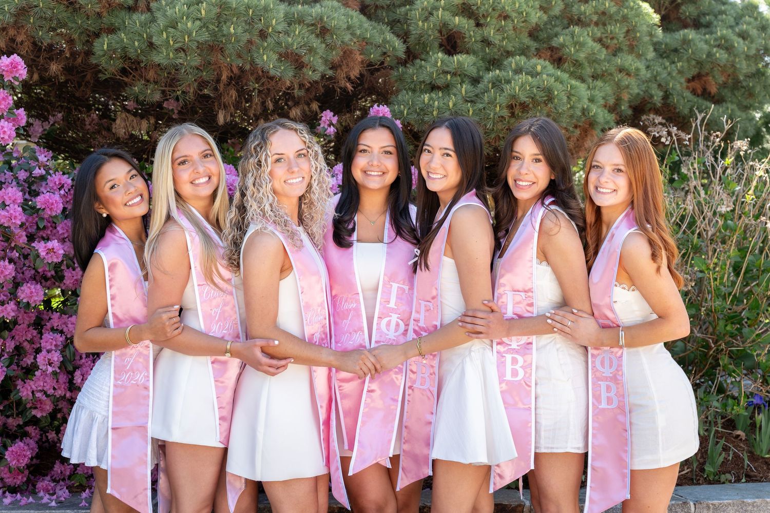 Seven young women in white dresses wearing pink sashes pose together outdoors near colorful flowers.